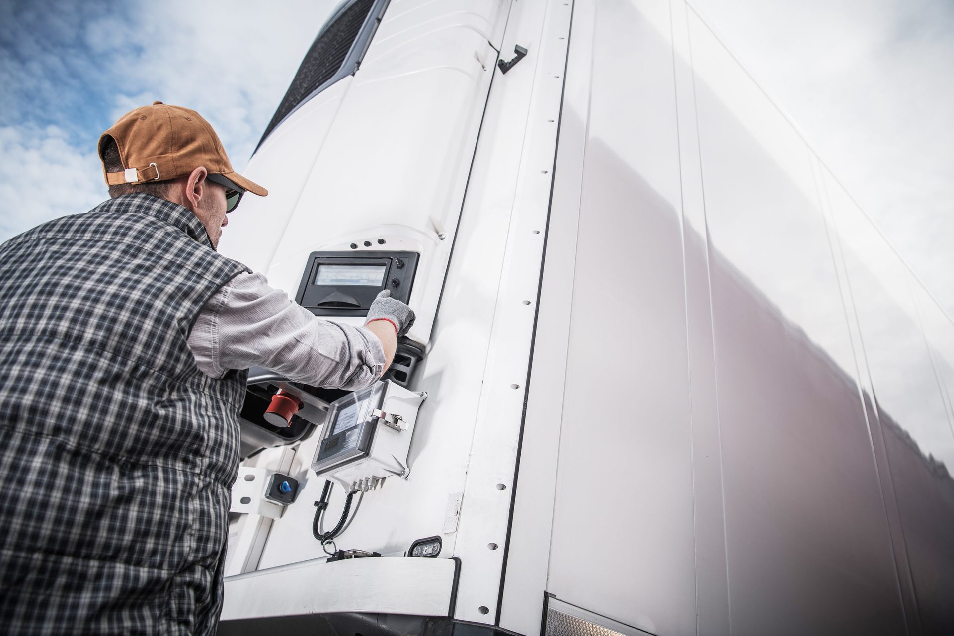 Man in plaid shirt and hat adjusting settings on a white refrigerated truck's control panel.