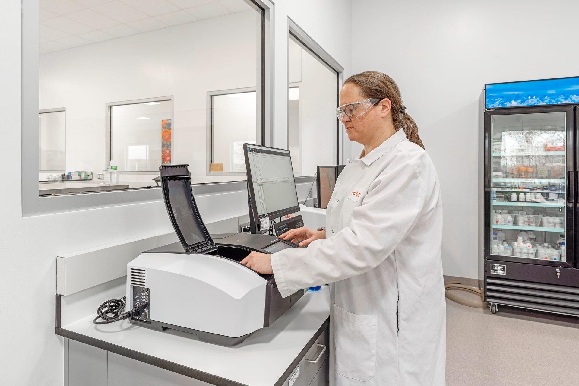 Researcher in safety glasses and lab coat operating an analytical instrument in a modern lab.