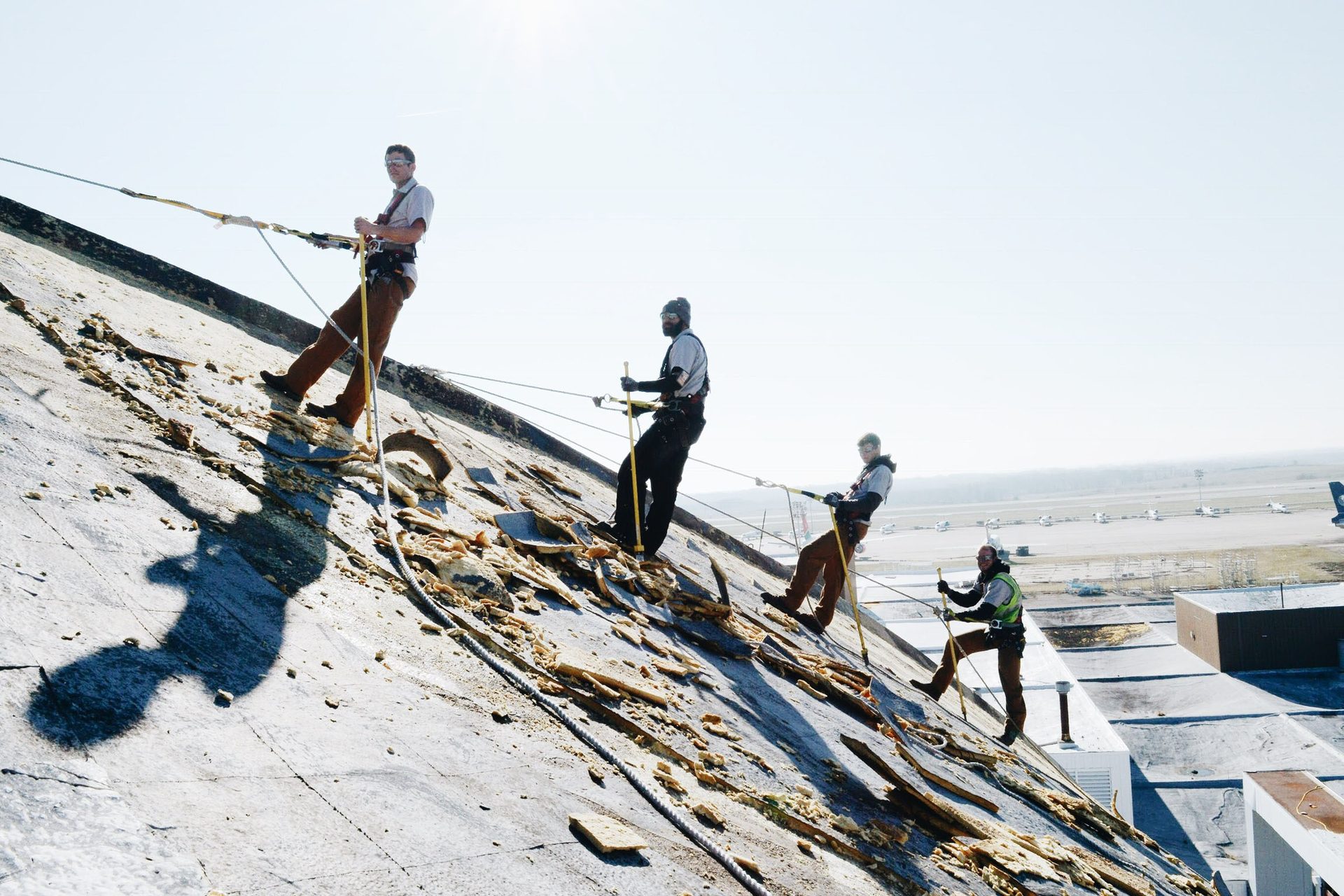Four workers rappel down a damaged roof, an airport in the background.