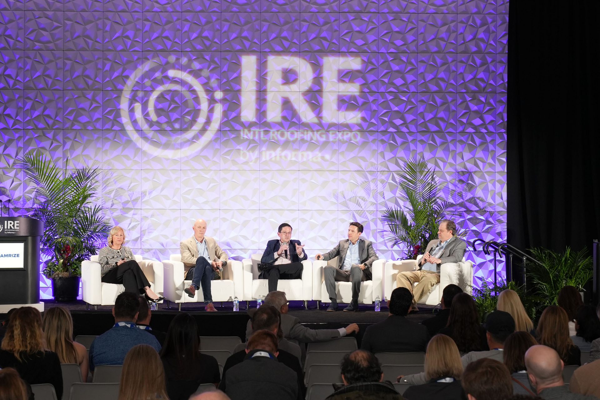 Five speakers on stage participate in a panel discussion at the IRE International Roofing Expo, with an audience watching.