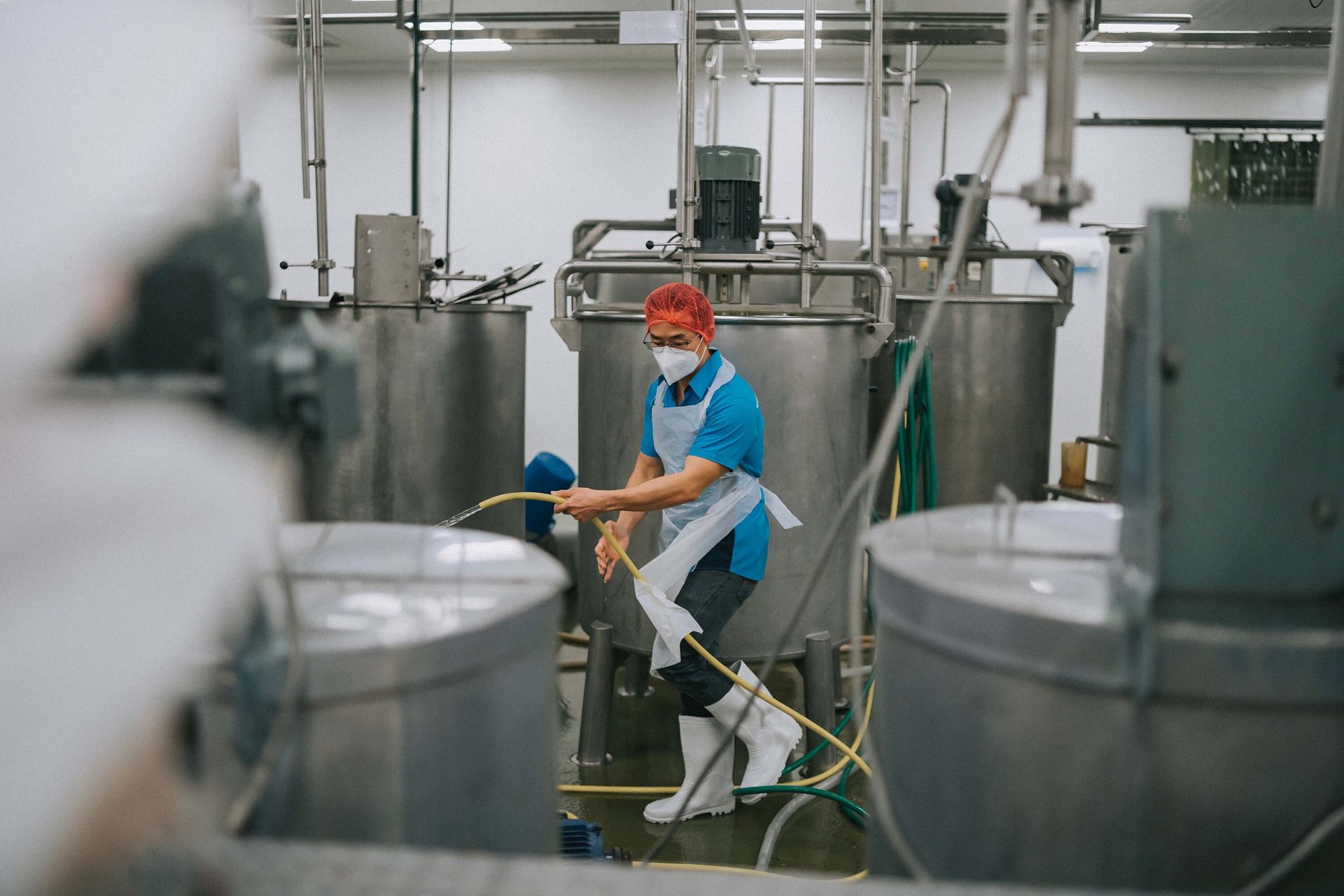 Worker in PPE cleaning a factory floor with a hose, surrounded by industrial tanks.