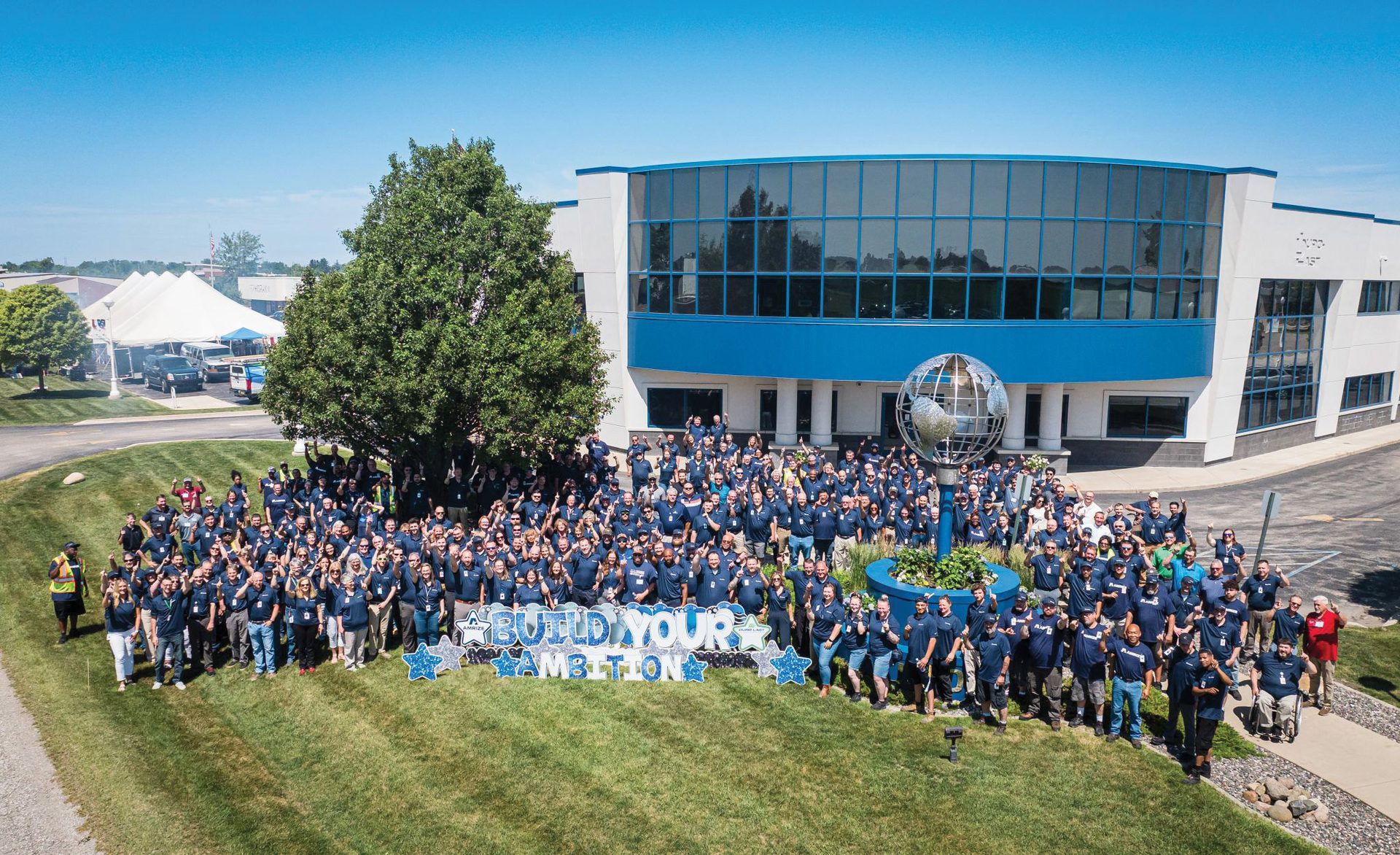 Aerial view: Large group of people in blue shirts pose outdoors by a modern building with an "AMBITION" sign.