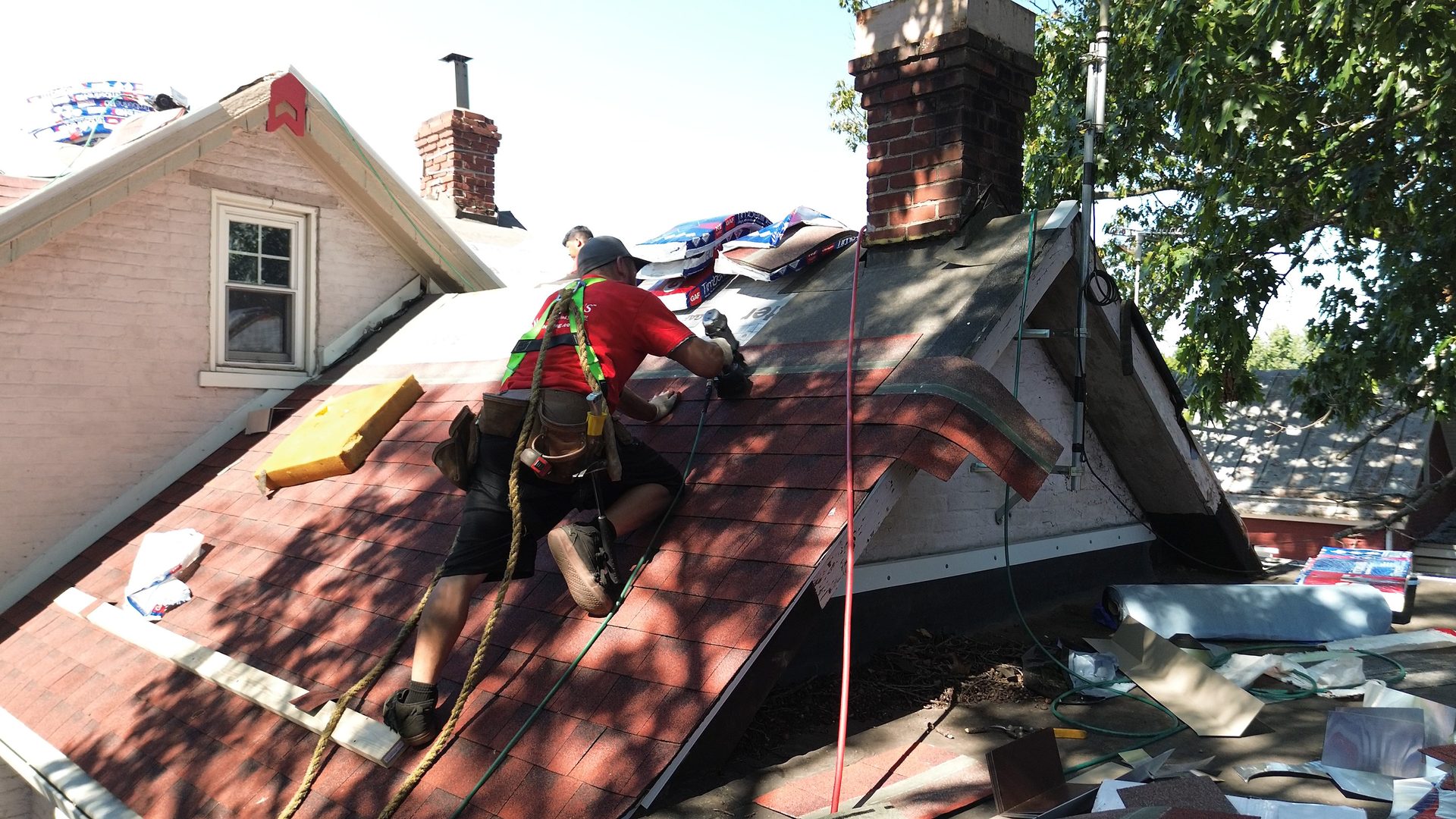 A roofer in a harness installs red shingles on a steep house roof, with materials nearby.