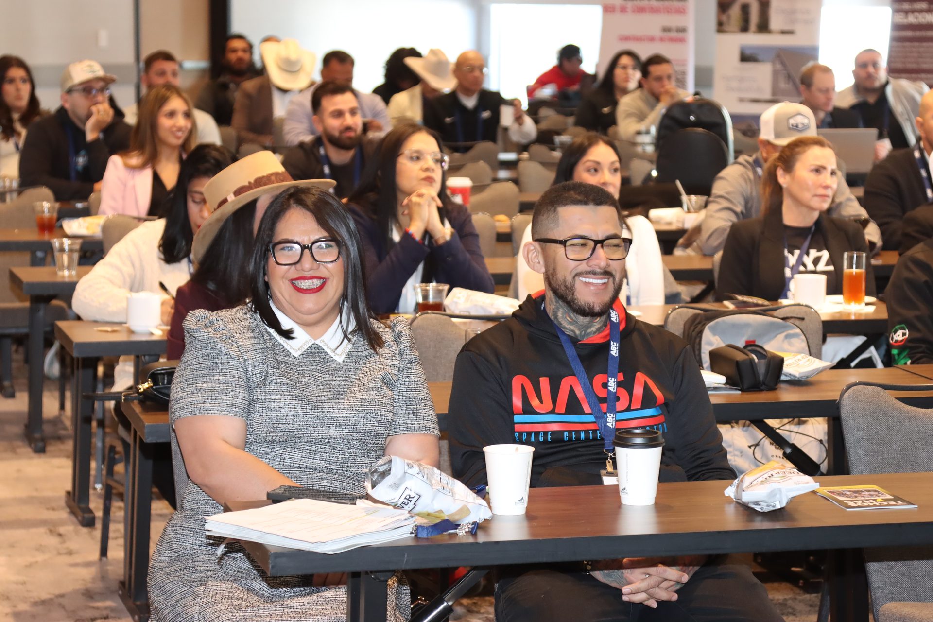 Smiling woman with braces and man in NASA hoodie at a conference with other attendees.