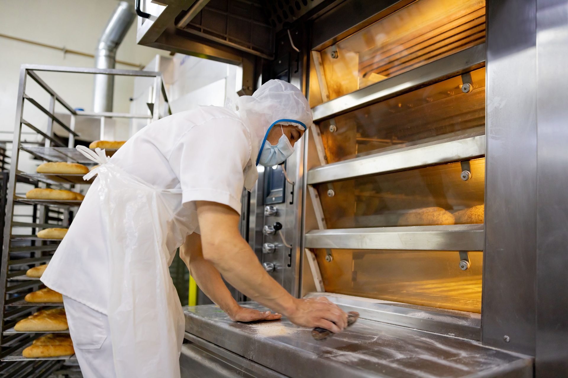 A baker in protective gear and mask cleans an industrial oven, with baked bread visible inside and on a rack.