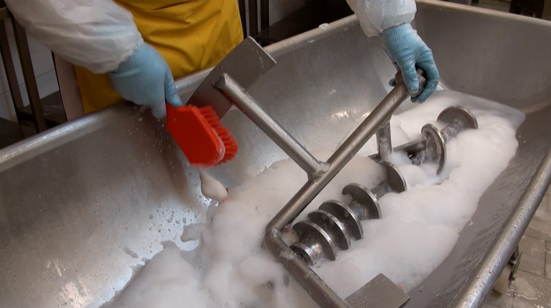 Hands in gloves cleaning large industrial mixer with brush and soap.