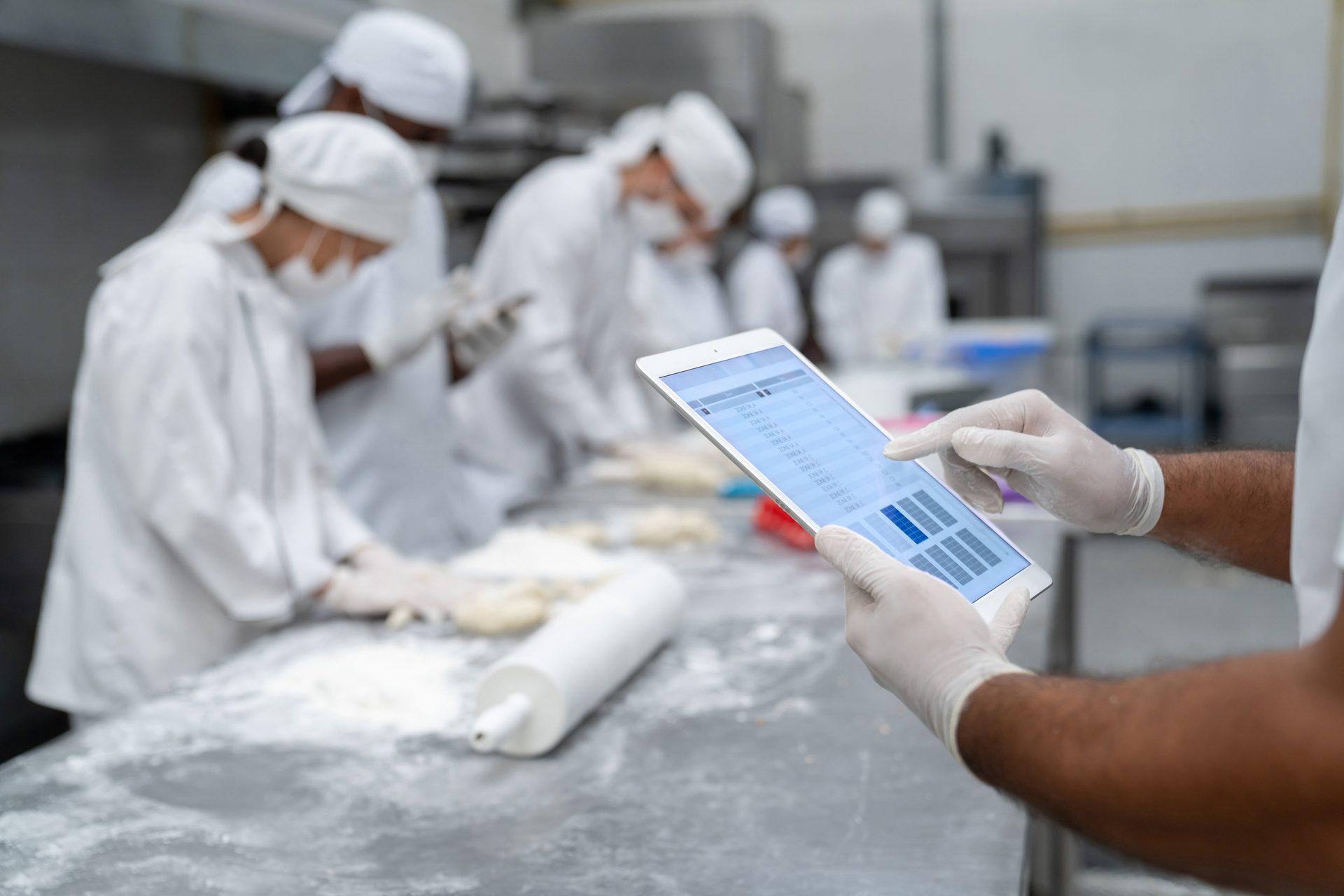 A gloved person uses a tablet to manage production in a busy bakery with masked workers kneading dough.