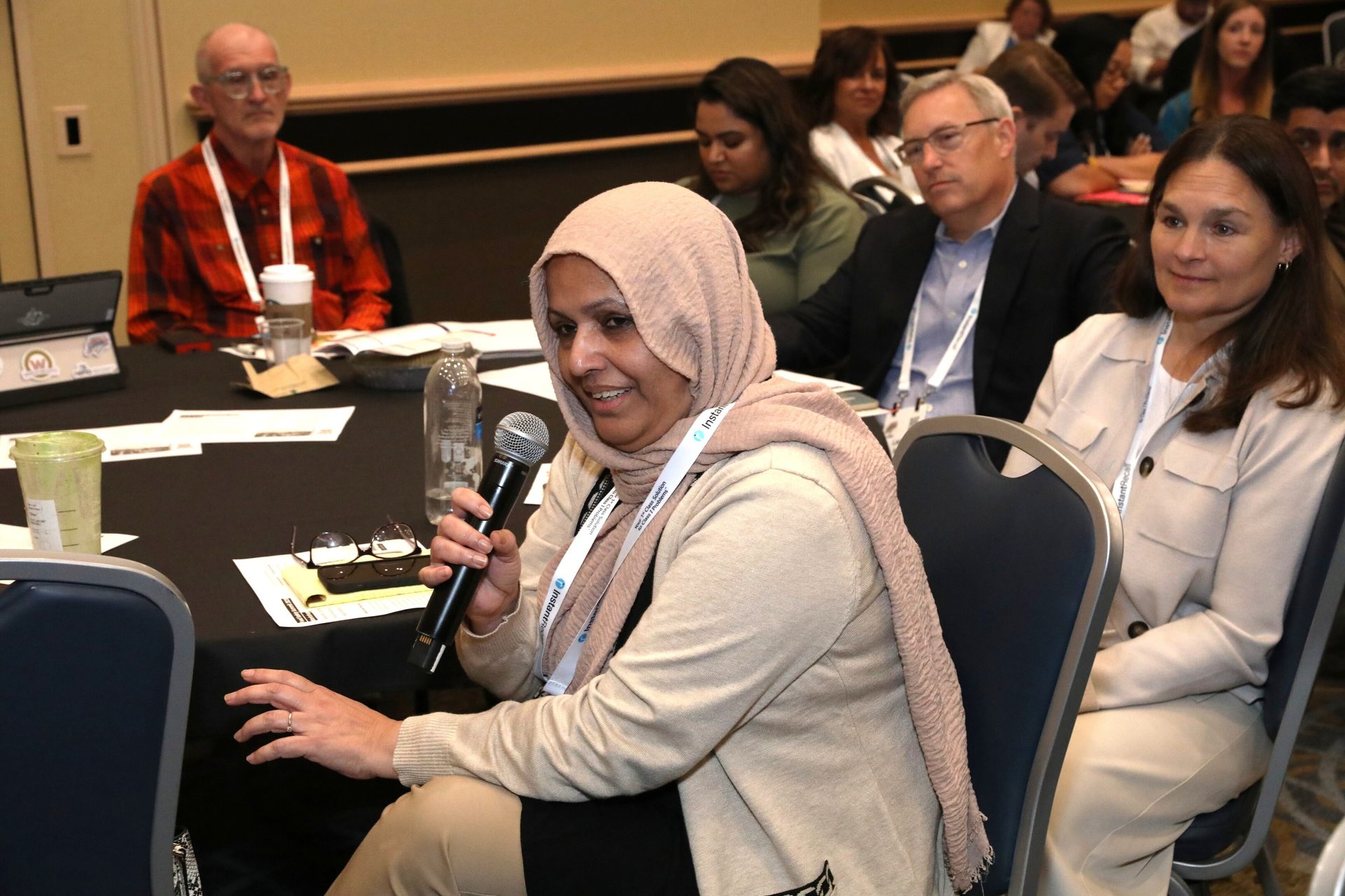 Woman in hijab speaking into a microphone at a conference, with other attendees.