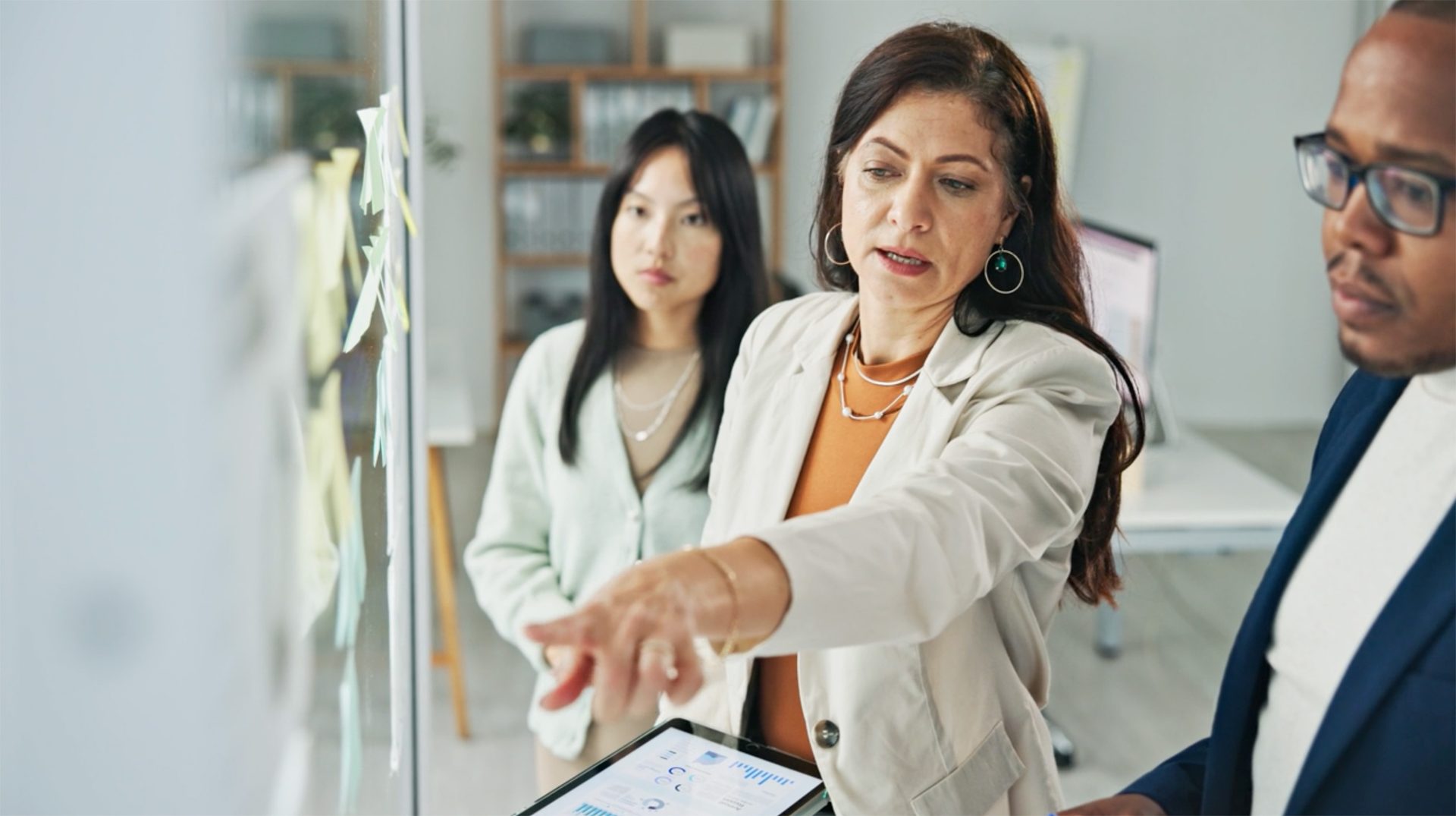 Business colleagues collaborating in office, woman pointing at tablet with data, other woman and man listening.