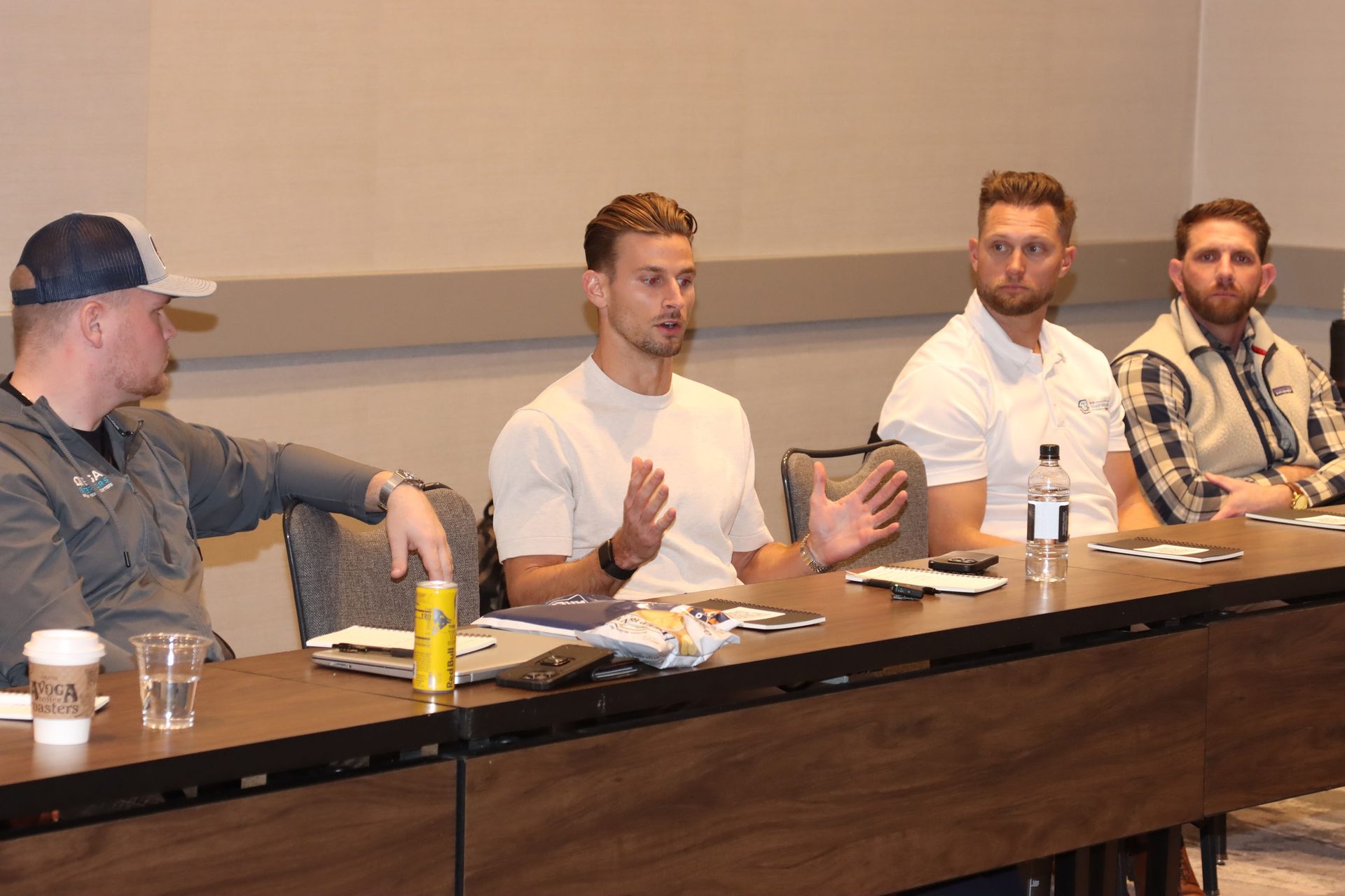 Four men seated at a long table during a meeting; one man actively speaks with hands raised.