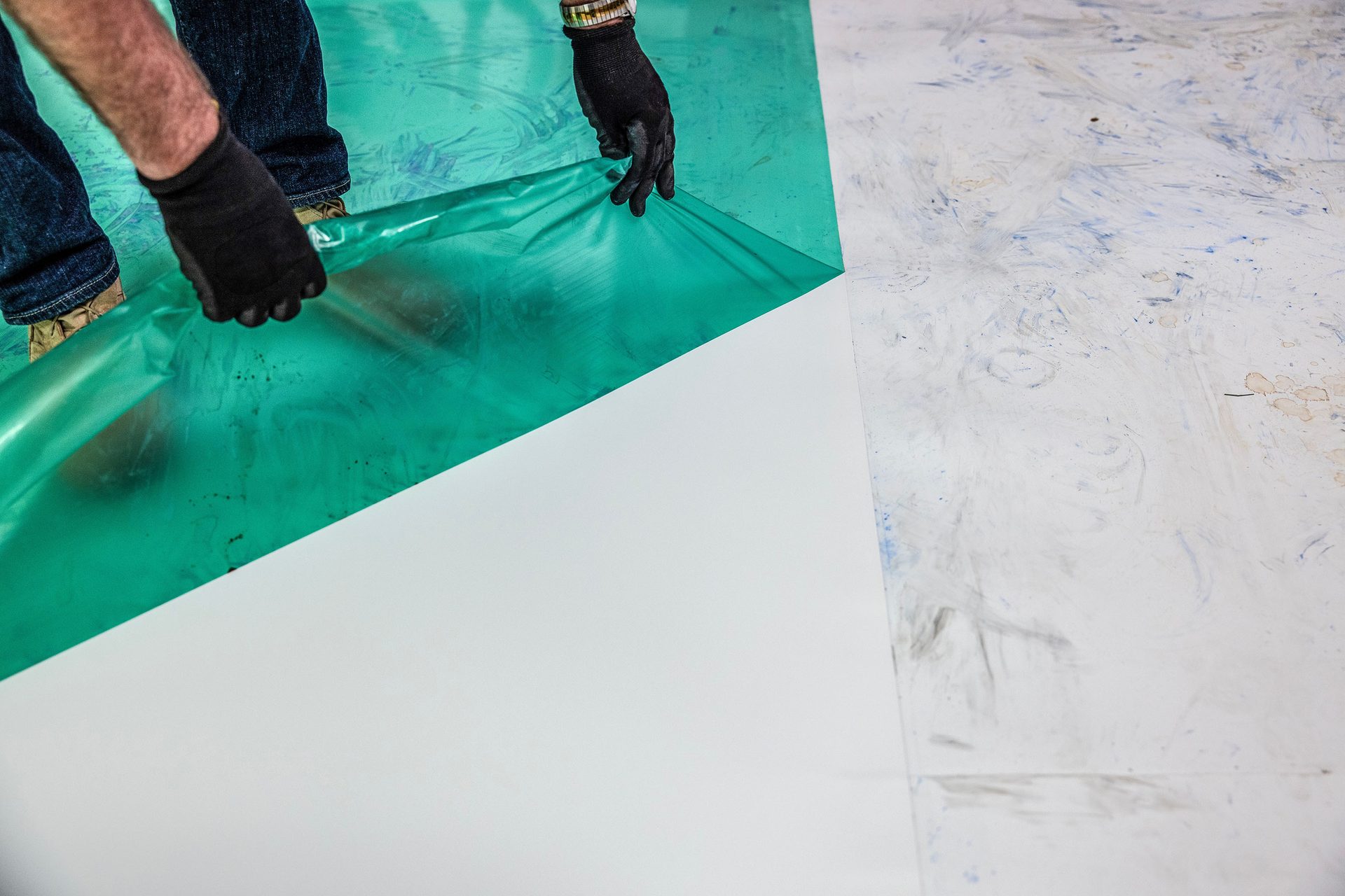Gloved hands peeling green film from a white surface, revealing its cleanliness against a dirty floor.