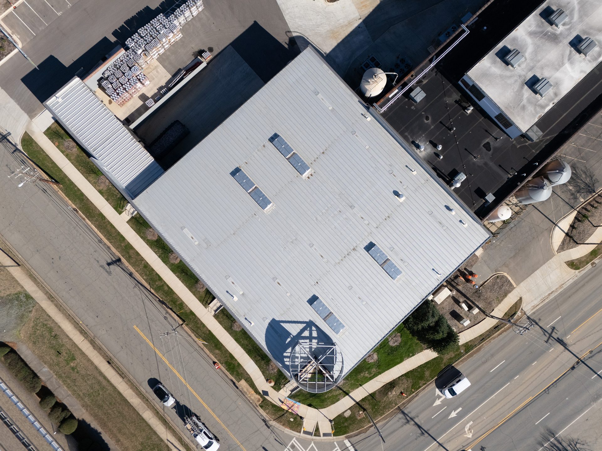 Aerial view of a large industrial building with a metal roof, roads, and surrounding structures.