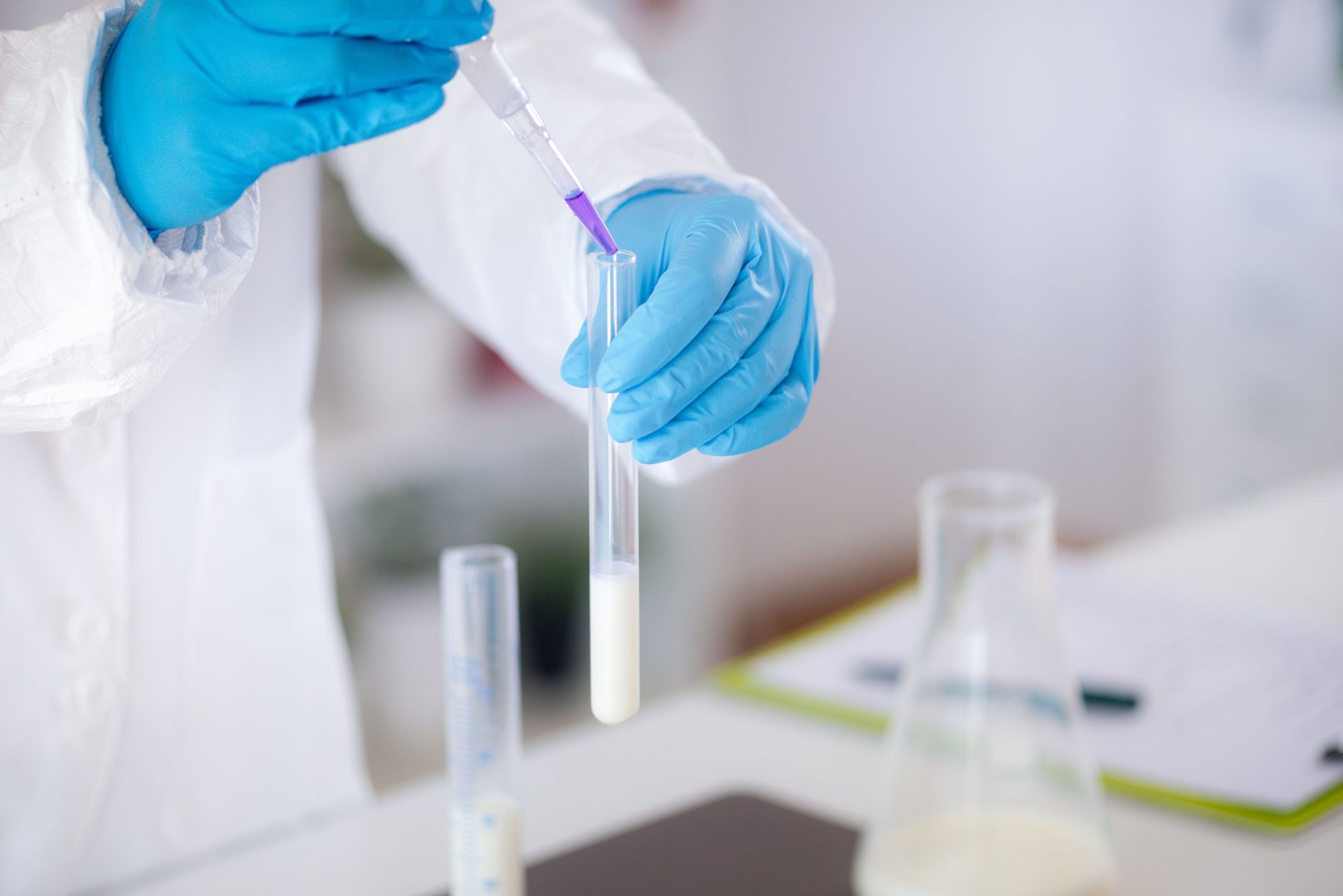 Hand in blue glove pipetting purple liquid into a test tube with white liquid in a lab.