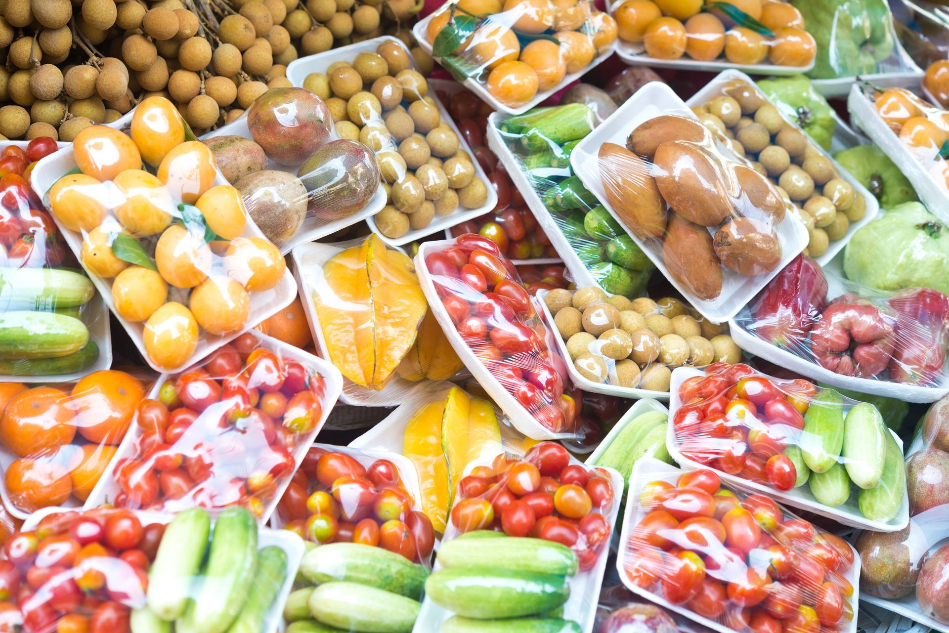 A vibrant market display of various fresh fruits and vegetables, neatly packaged in clear plastic wrap on white foam trays.