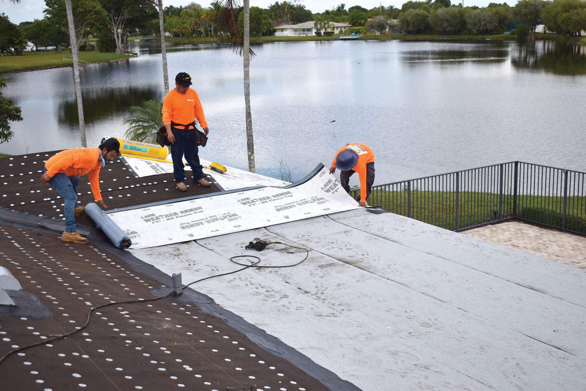 Three roofers install weather-armor roofing material on a house overlooking a scenic lake.