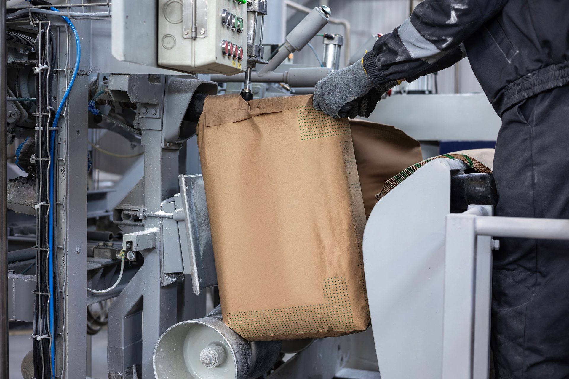 Worker's gloved hands operating industrial machinery to fill a large brown paper bag with material.
