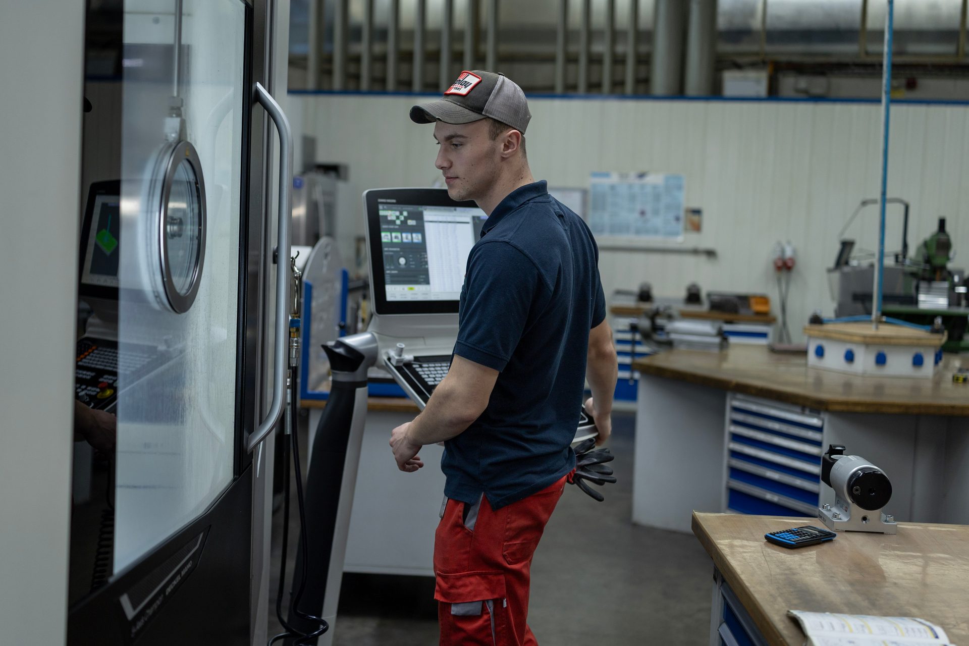 Young man in a cap operating a CNC machine in a workshop, checking the control panel.