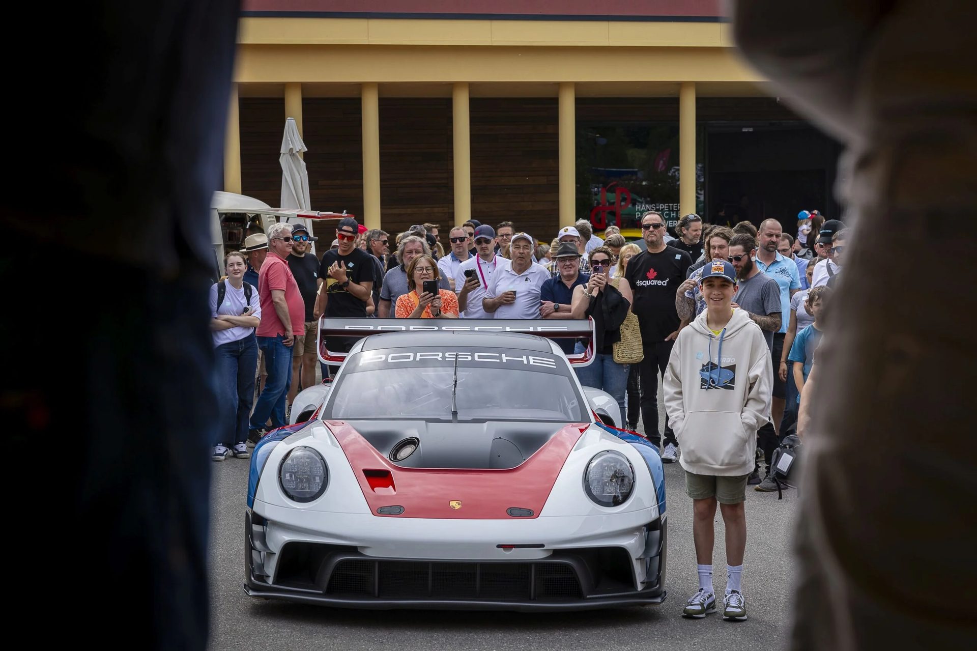 A custom Porsche race car with a red, white, and blue livery is on display, surrounded by a crowd.