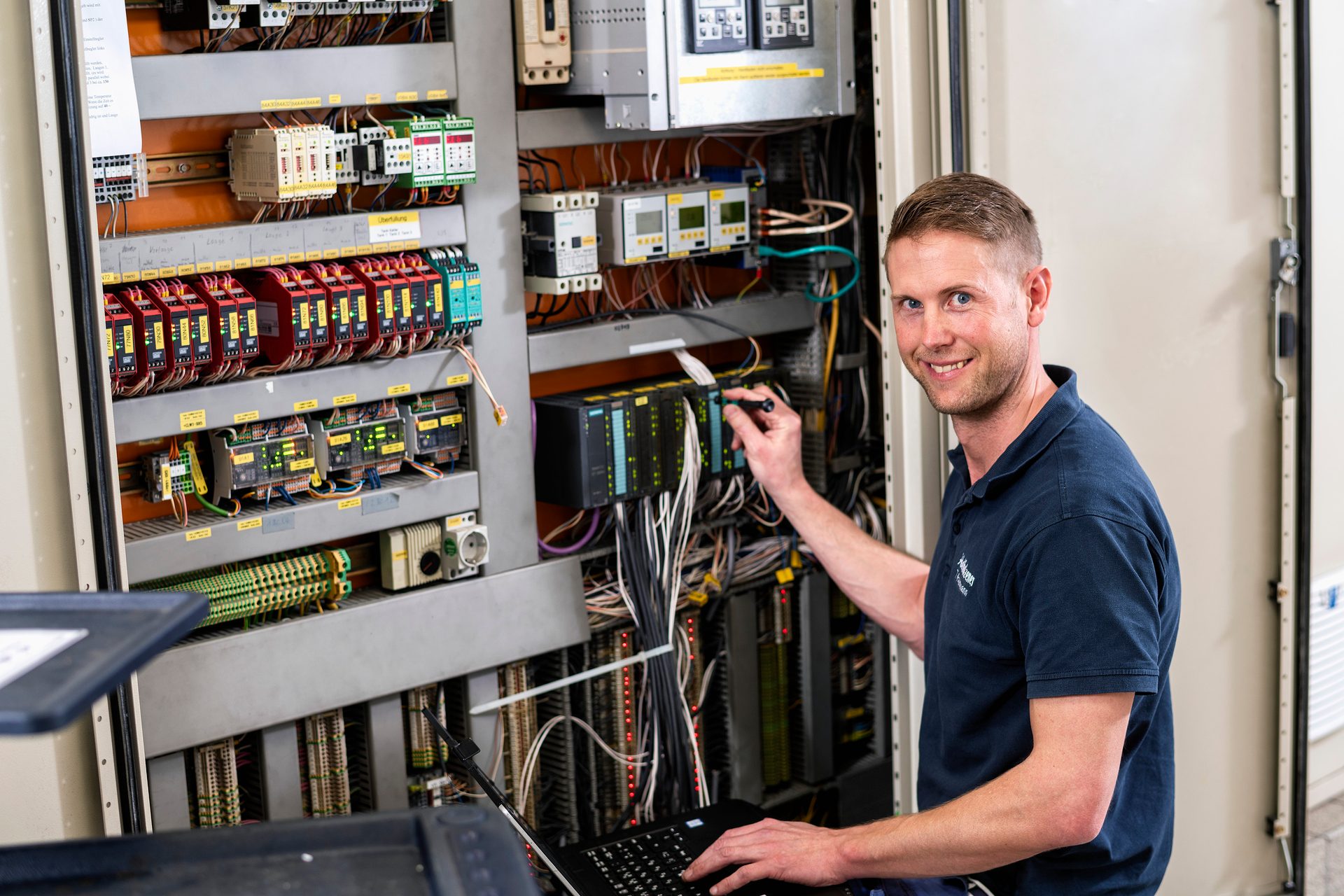 Smiling technician debugging an electrical control panel with a laptop.