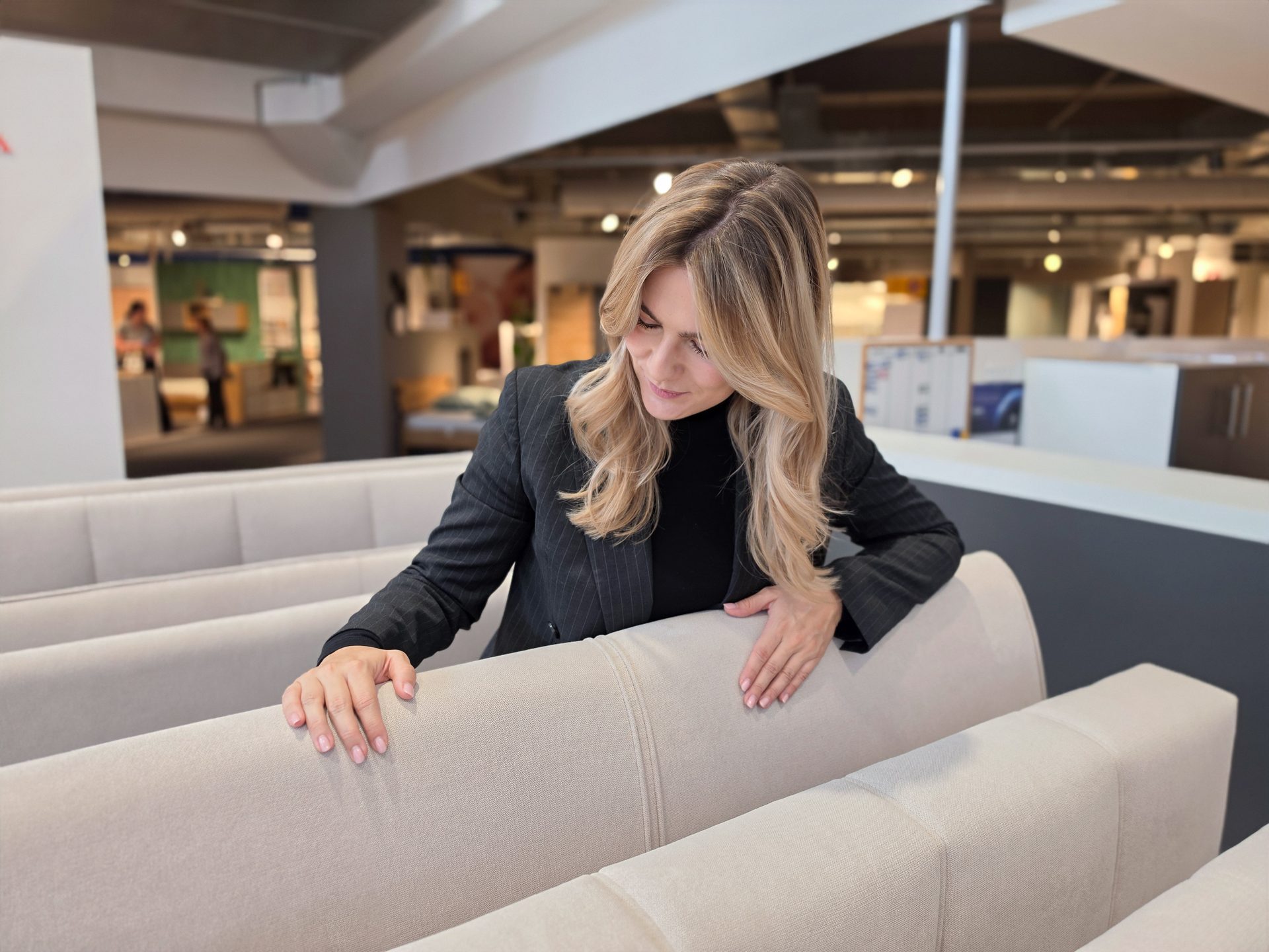 Woman in black suit jacket touching a beige sofa in a furniture store, looking down.