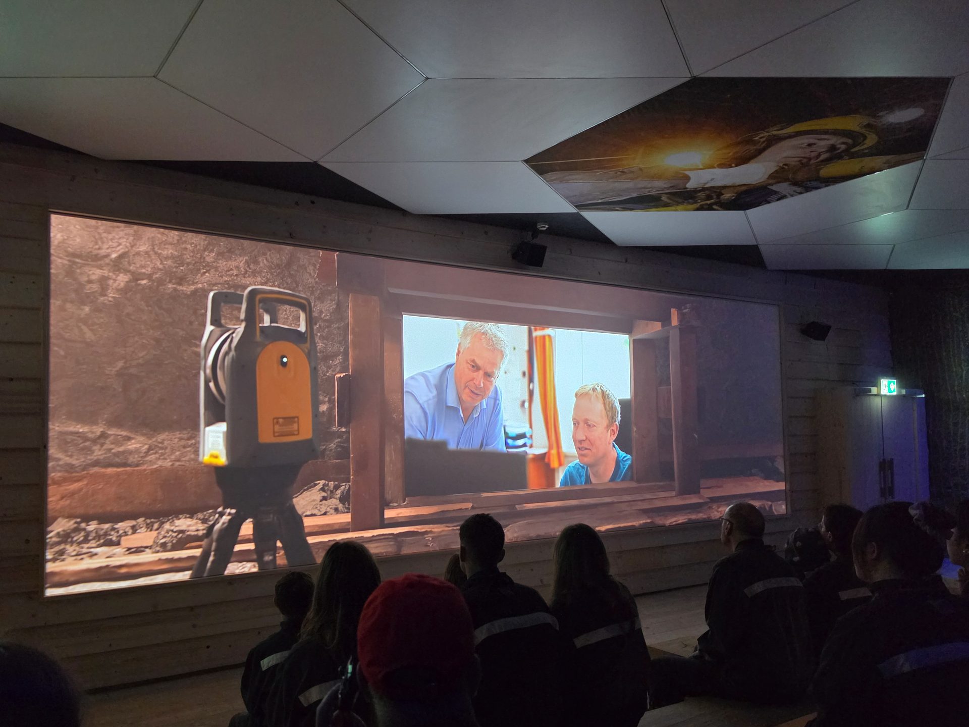 People sit in a dark room watching a large screen displaying two men and a surveying instrument.