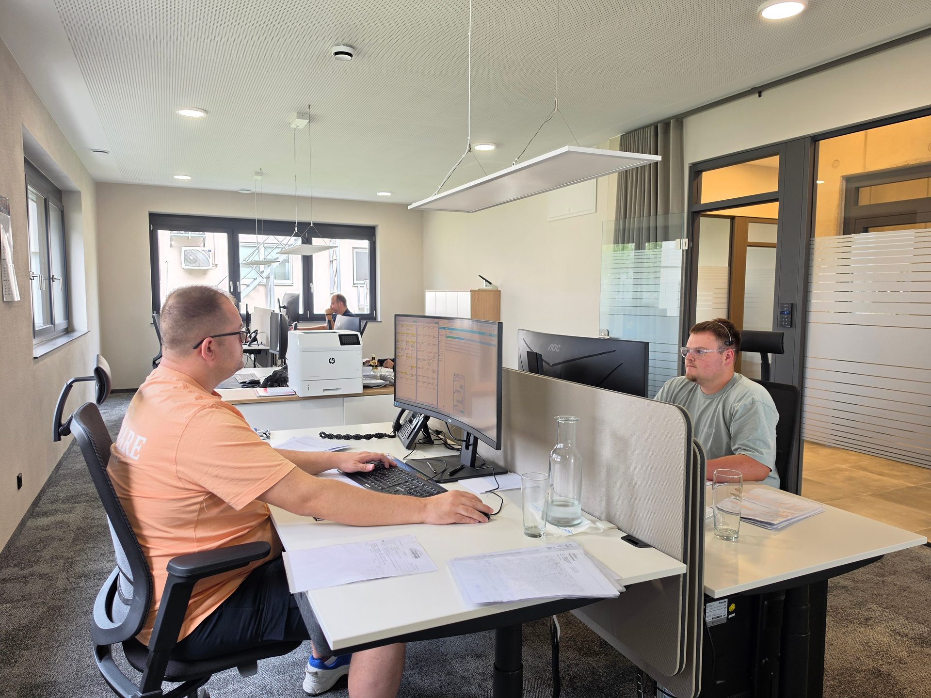 Two men work at their desks in a modern office, one in an orange shirt and one in a green shirt.