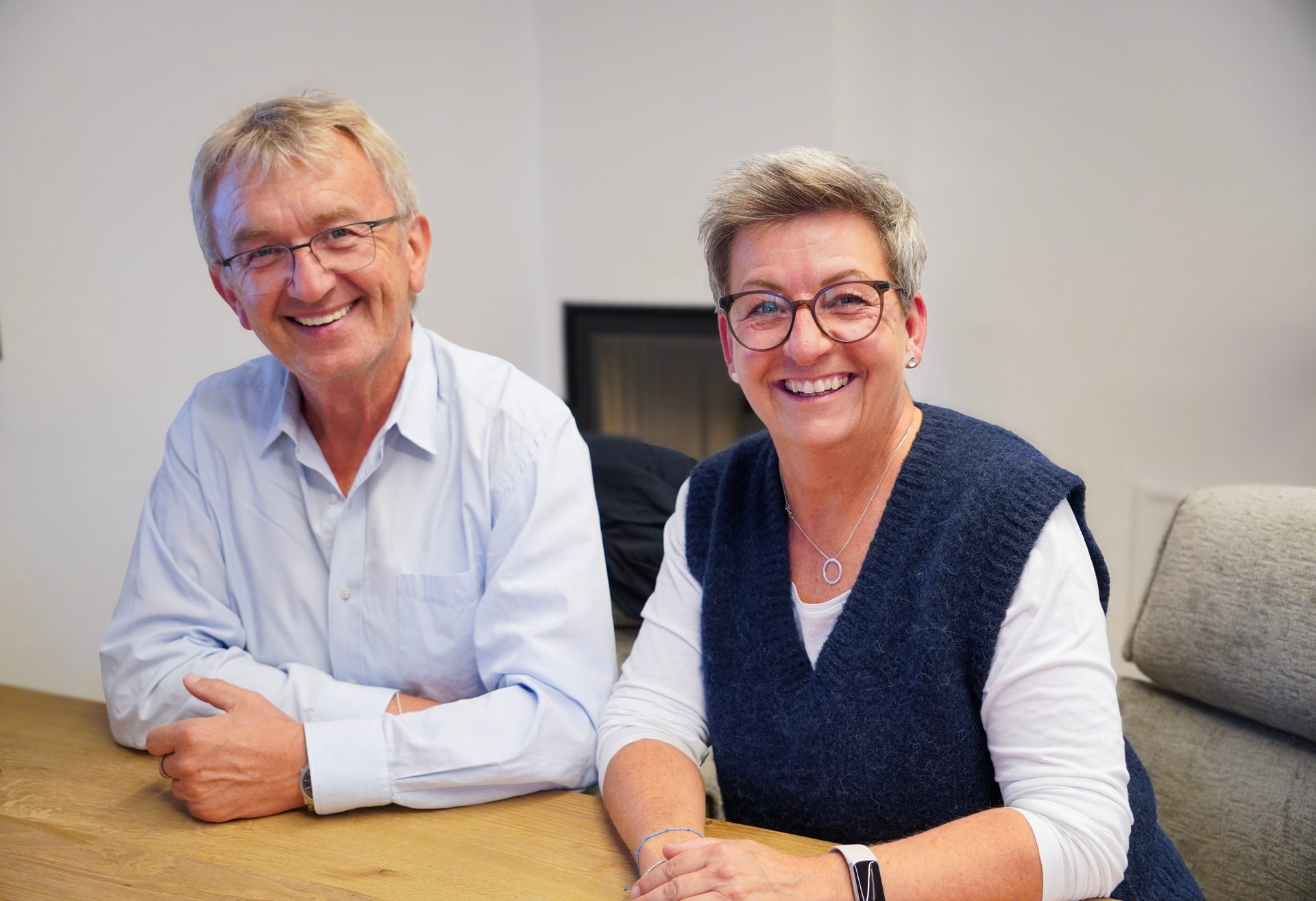 Two smiling people, a man and a woman, seated at a wooden table, looking directly at the camera.