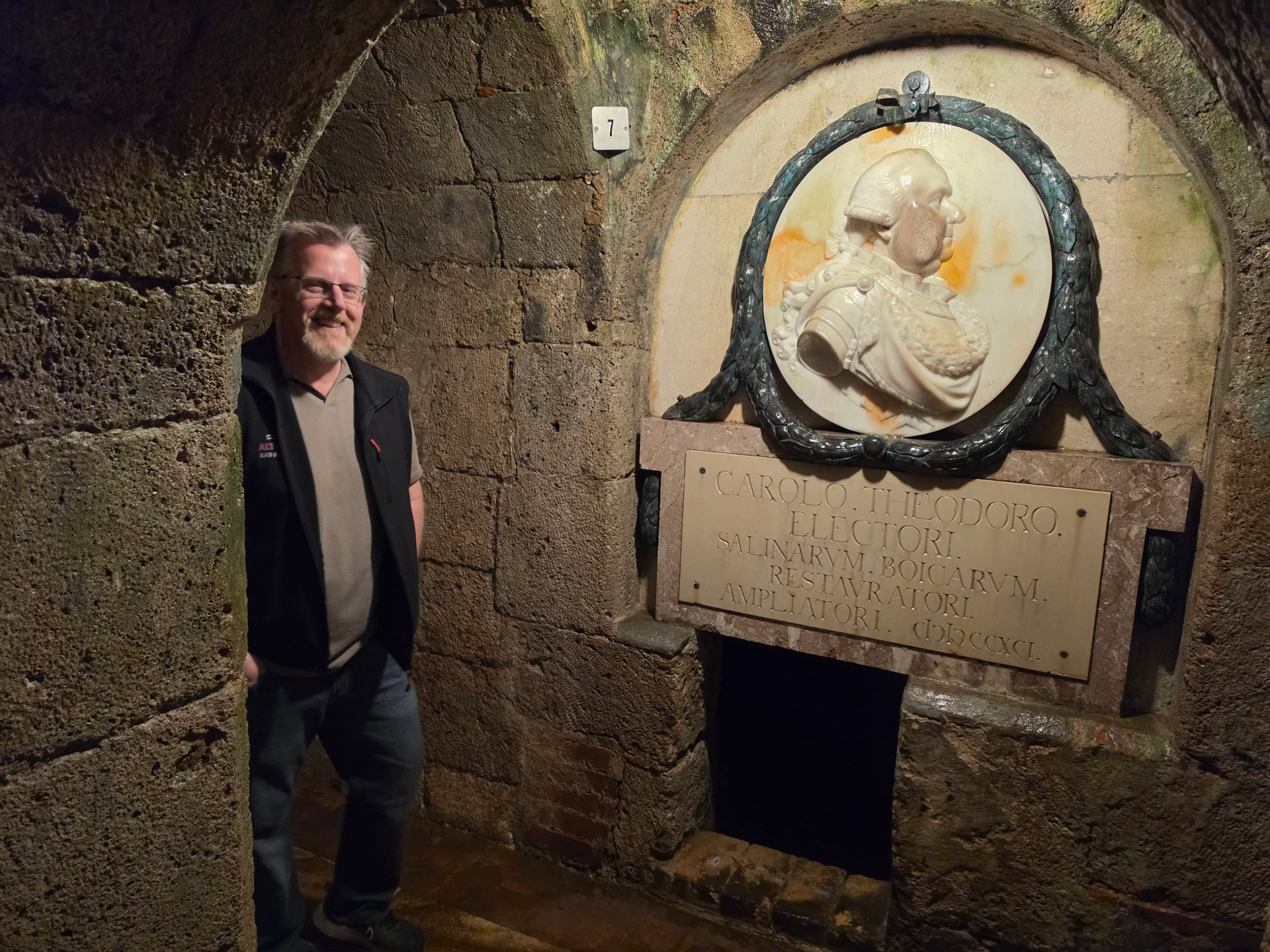 A smiling man stands in a stone archway next to a historical relief and plaque with Latin text.