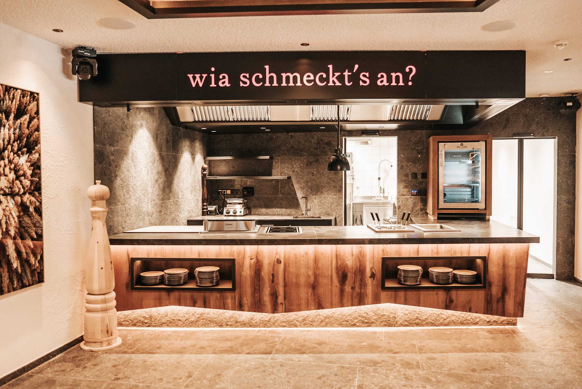 Modern open kitchen island with cooking equipment, glowing sign "wia schmeckt's an?", and wooden base.