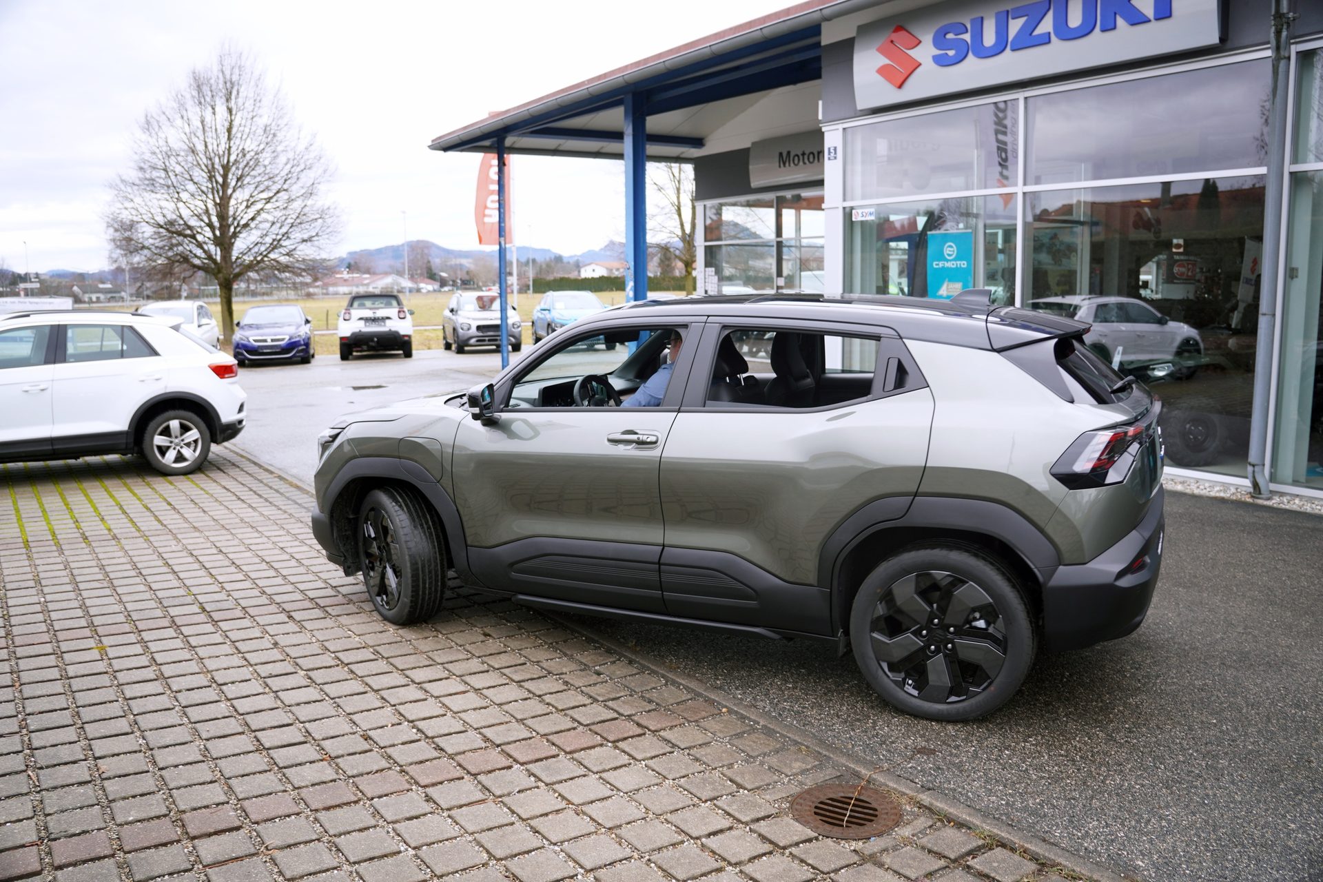 A military green SUV with black accents parked on pavers outside a Suzuki dealership on an overcast day.