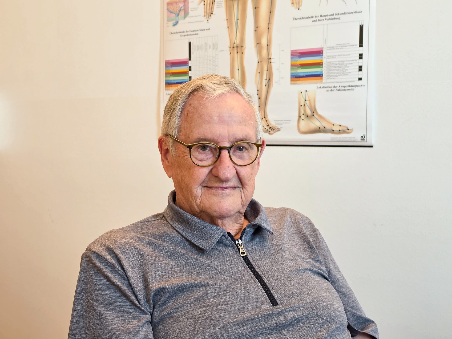 An elderly man with glasses and a gray collared shirt smiles at the camera, an acupuncture chart visible behind him.