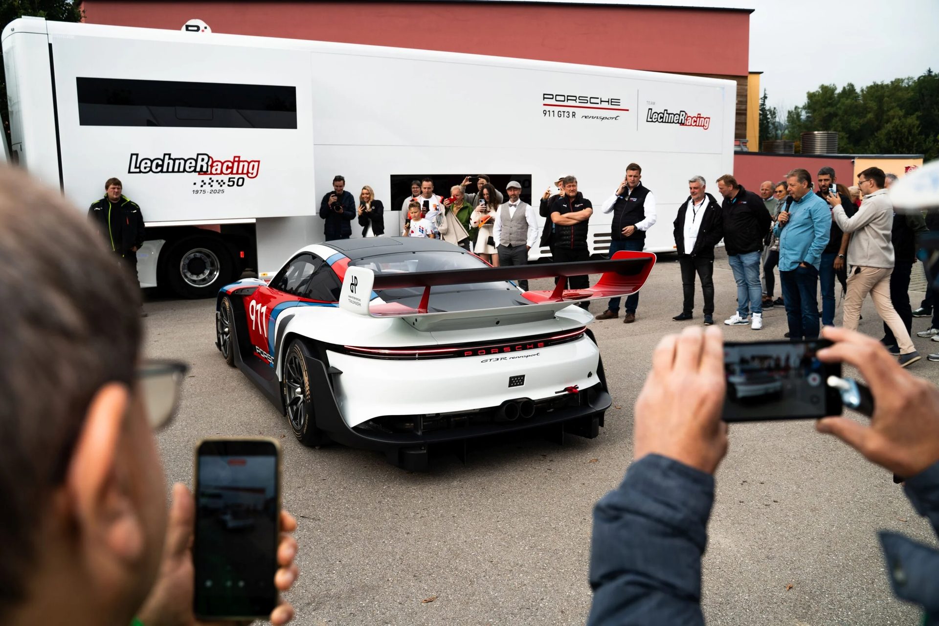 A Porsche 911 GT3 R Rennsport in red, white, and blue, surrounded by spectators and a Lechner Racing trailer.
