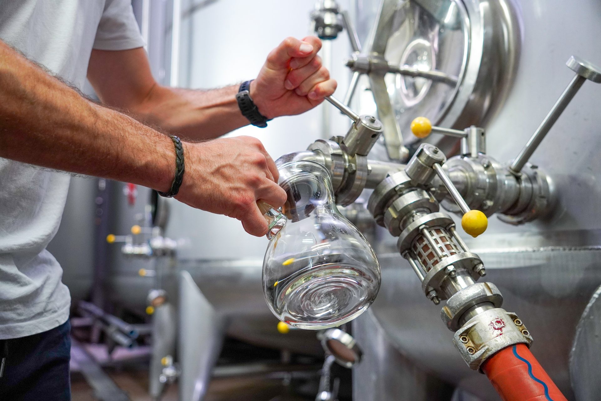 A person's hands holding a glass flask while operating industrial stainless steel brewing equipment.