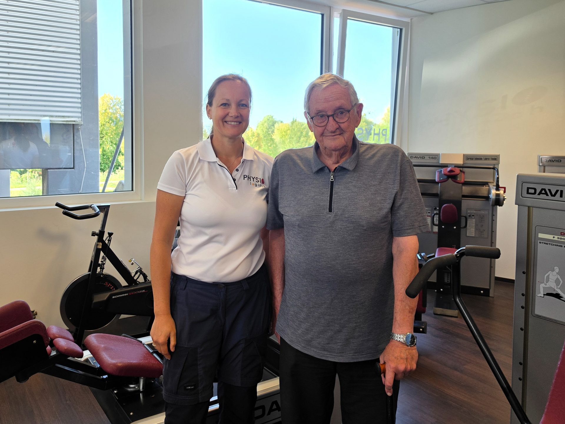 A physiotherapist and an older male patient smile while standing in a modern gym with exercise equipment.