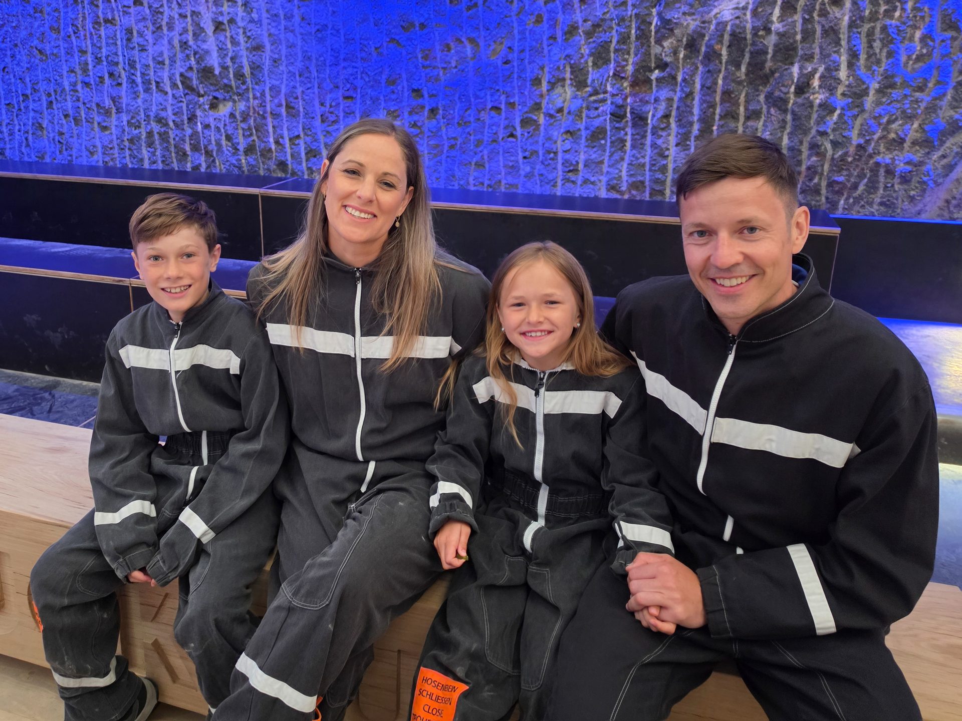 A smiling family of four, wearing matching black and white jumpsuits, sits in front of a blue textured wall.