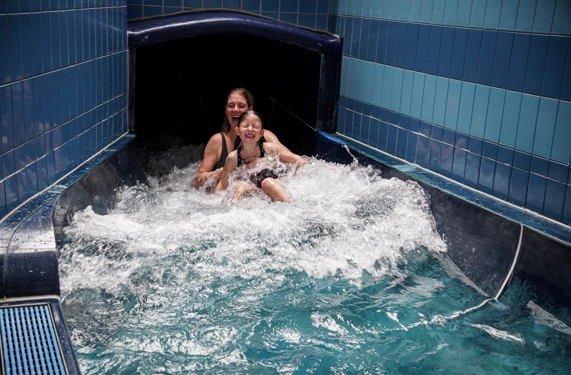 A woman and a young girl laughing joyfully as they splash down a blue water slide in a pool.