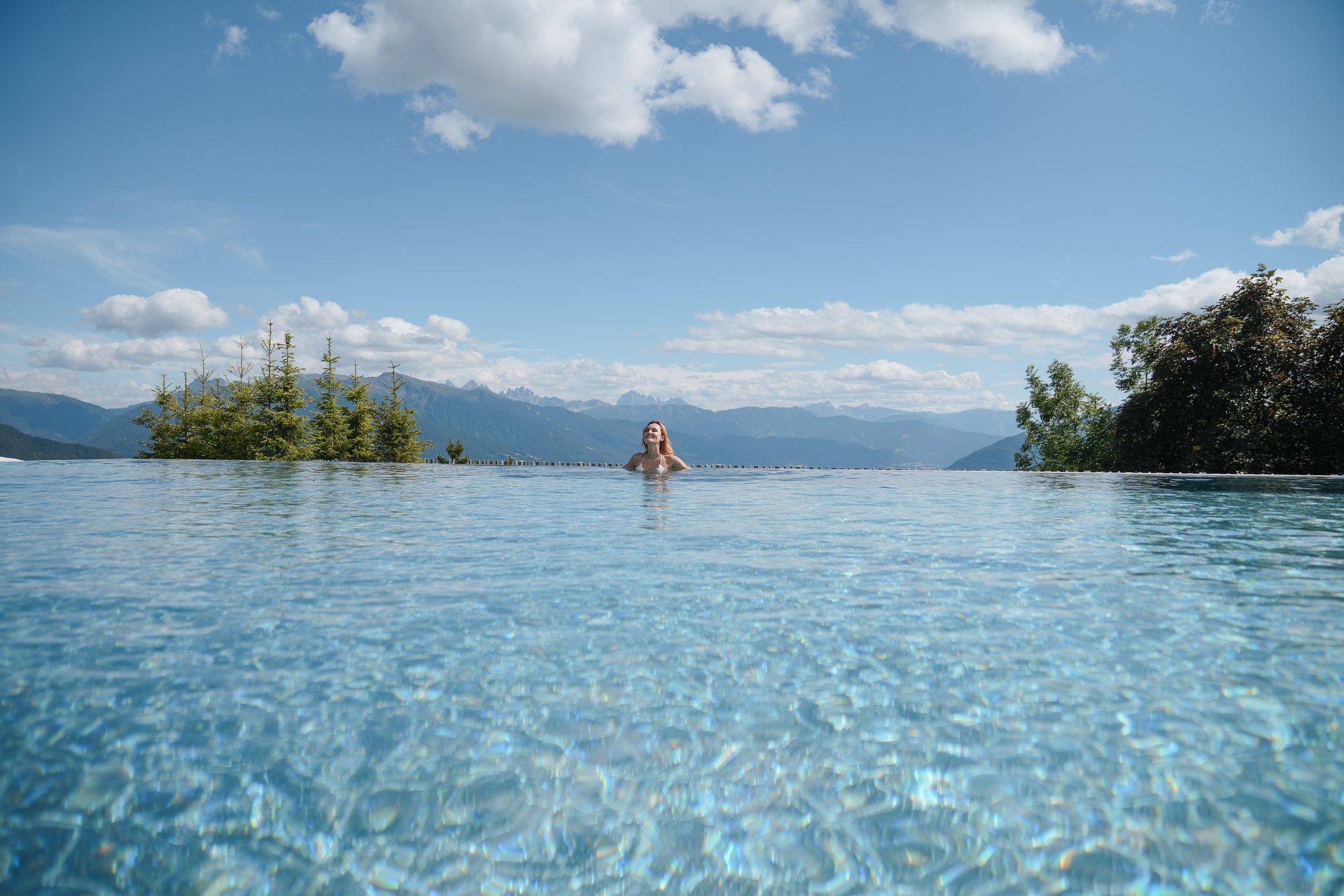 A woman relaxes in an infinity pool with a panoramic view of mountains under a blue sky.