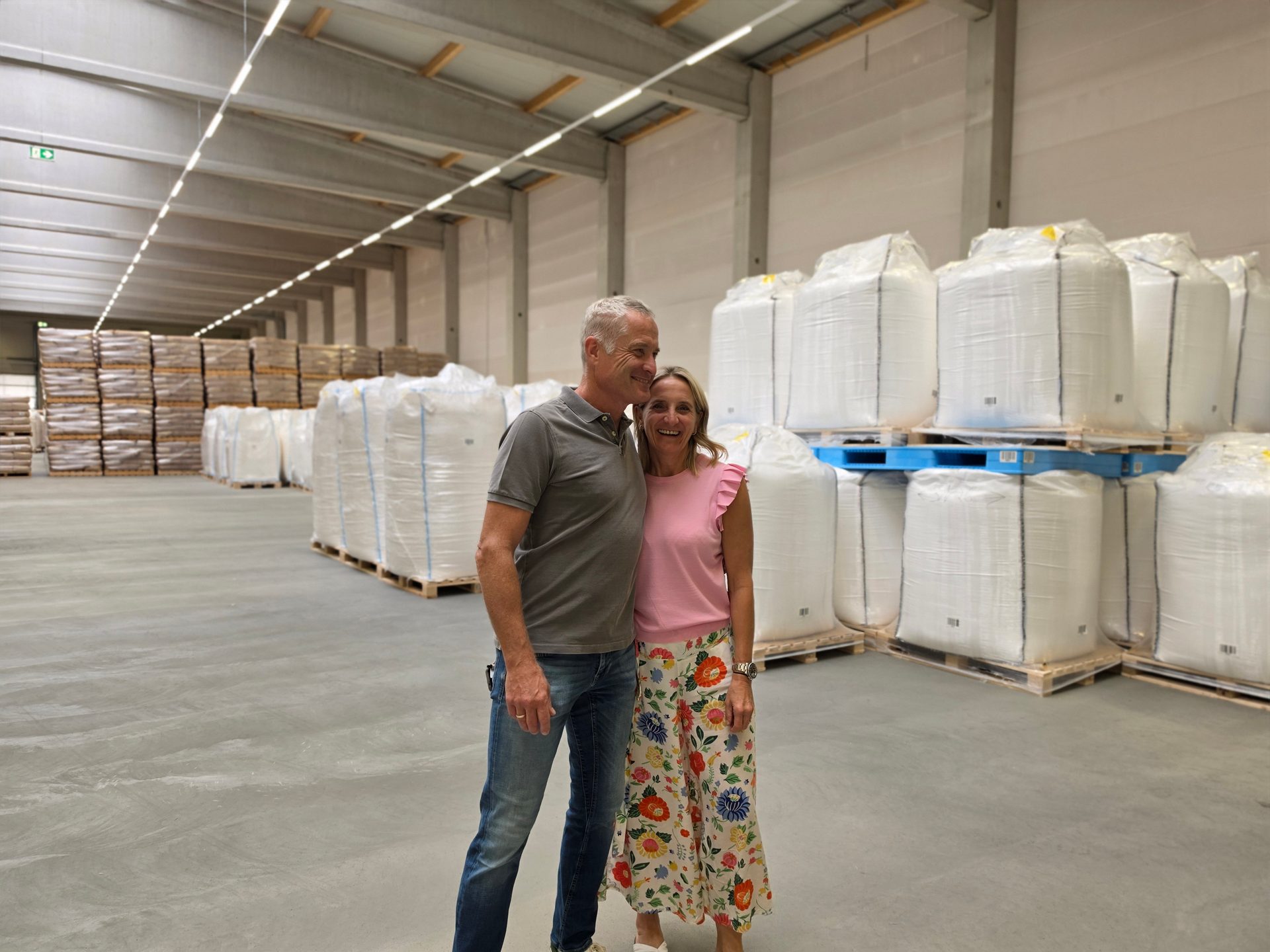 A smiling couple standing in a large warehouse filled with stacked white bulk bags and wooden pallets.
