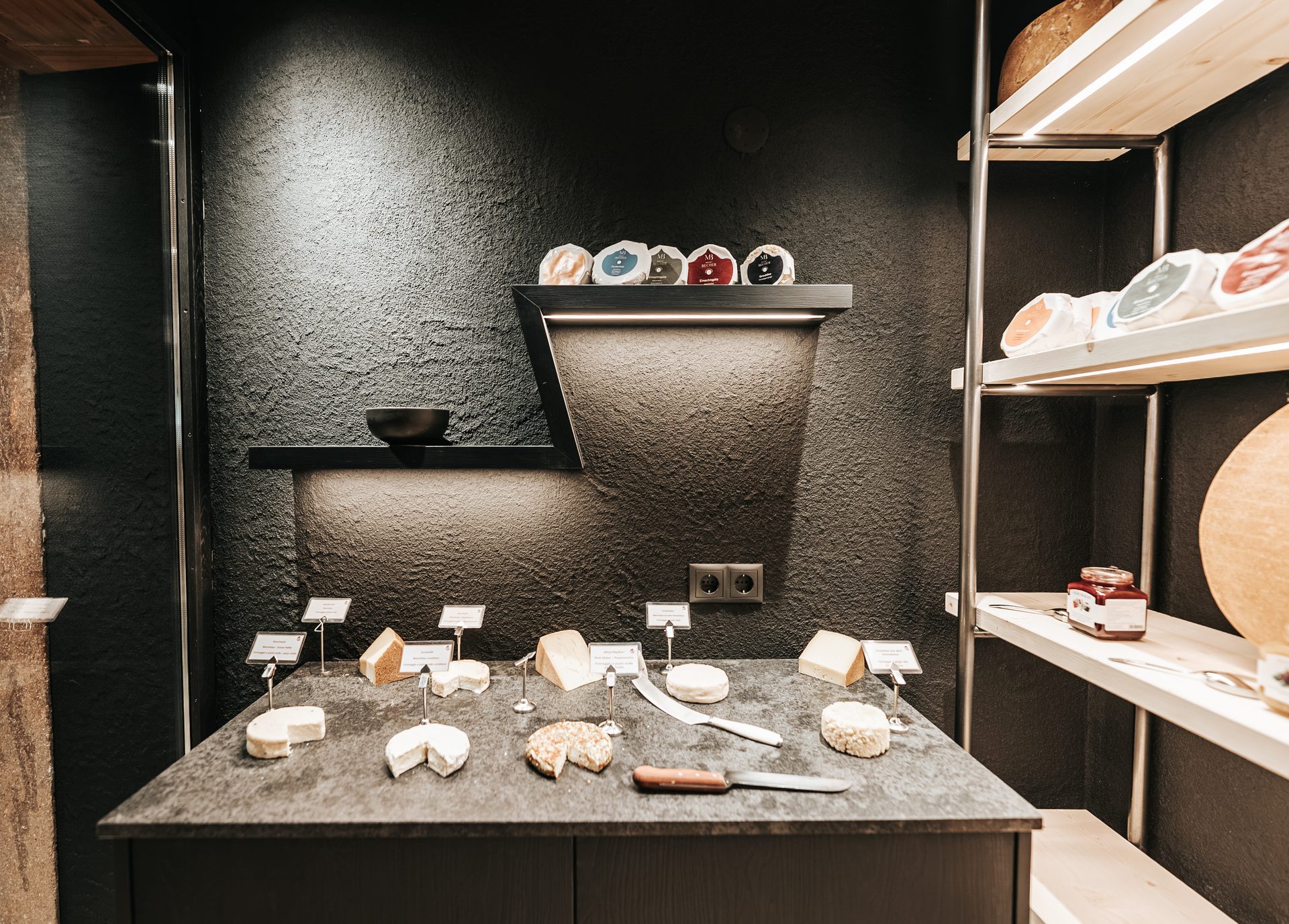 A dark-themed cheese shop with various artisanal cheeses displayed on a counter and shelves, with labels.