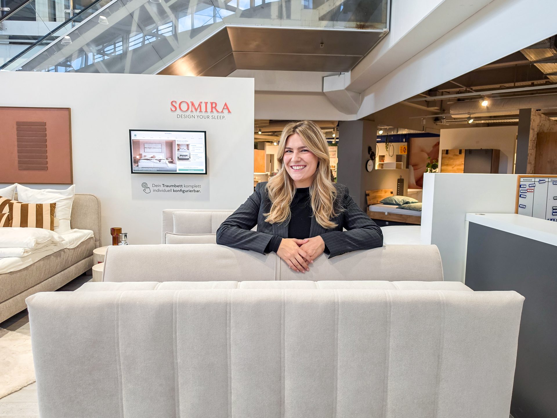 A smiling woman in a business suit leans on a sofa in a modern furniture store with Somira branding.