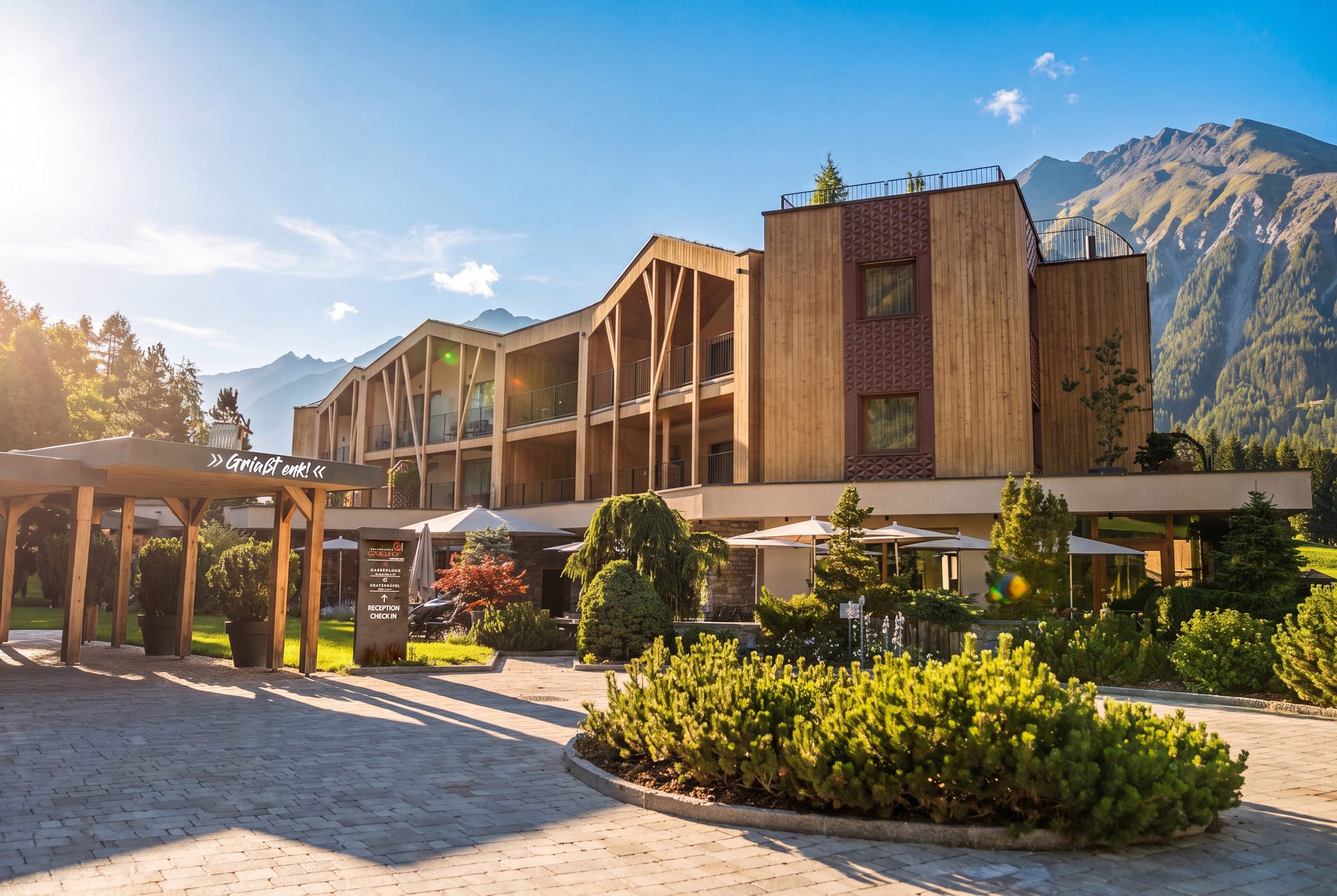 Modern mountain resort hotel with a wooden facade, balconies, and a spacious entrance garden under a clear sky.
