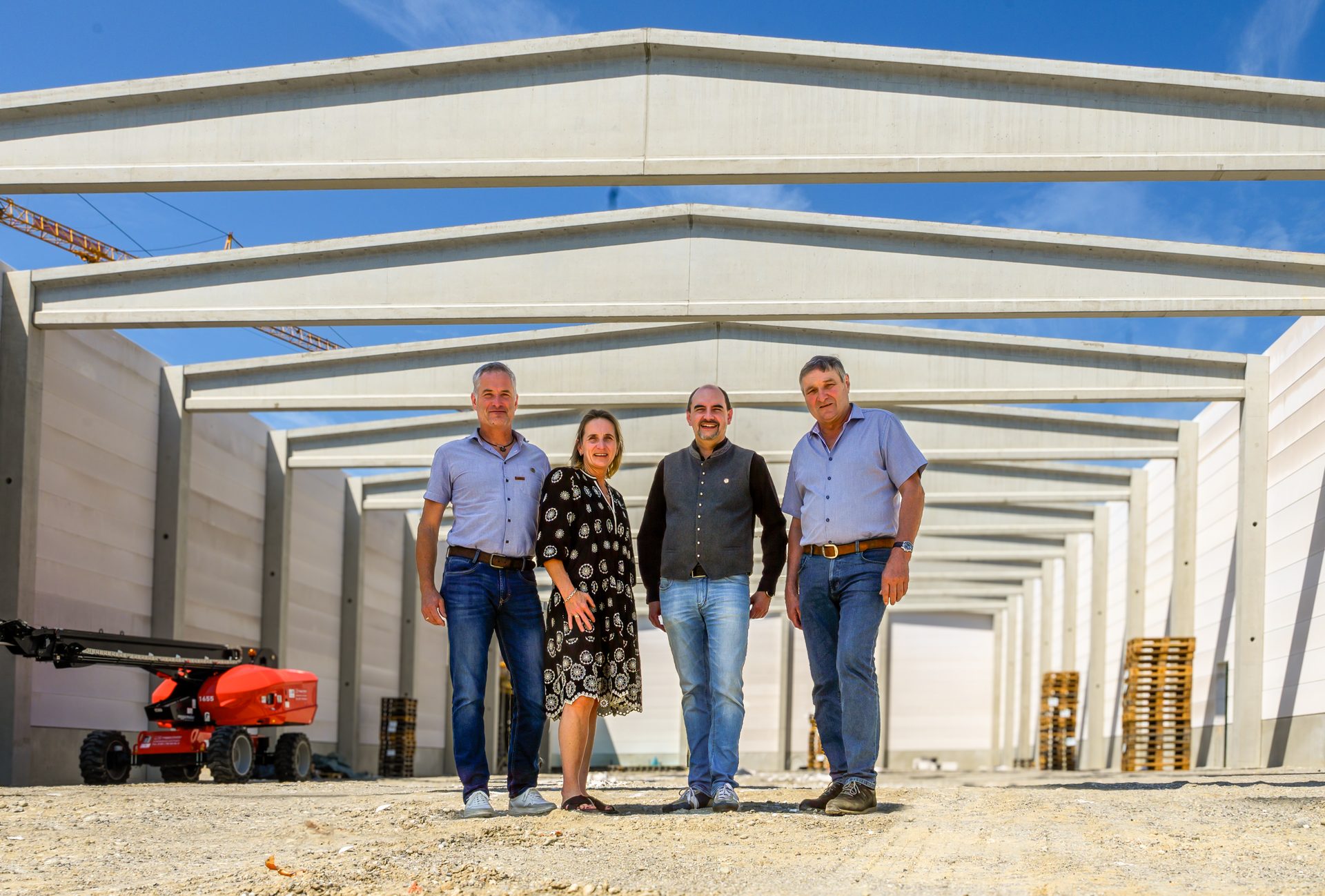 Four people stand in front of a large concrete building frame under construction on a sunny day.