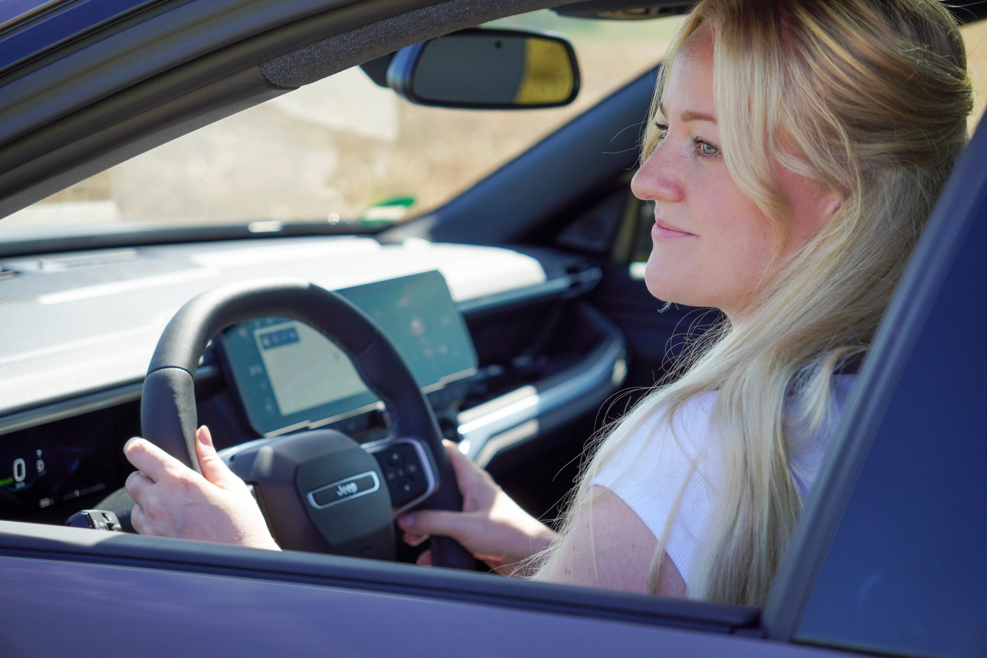 Blonde woman driving a modern car, hands on the steering wheel, looking forward.