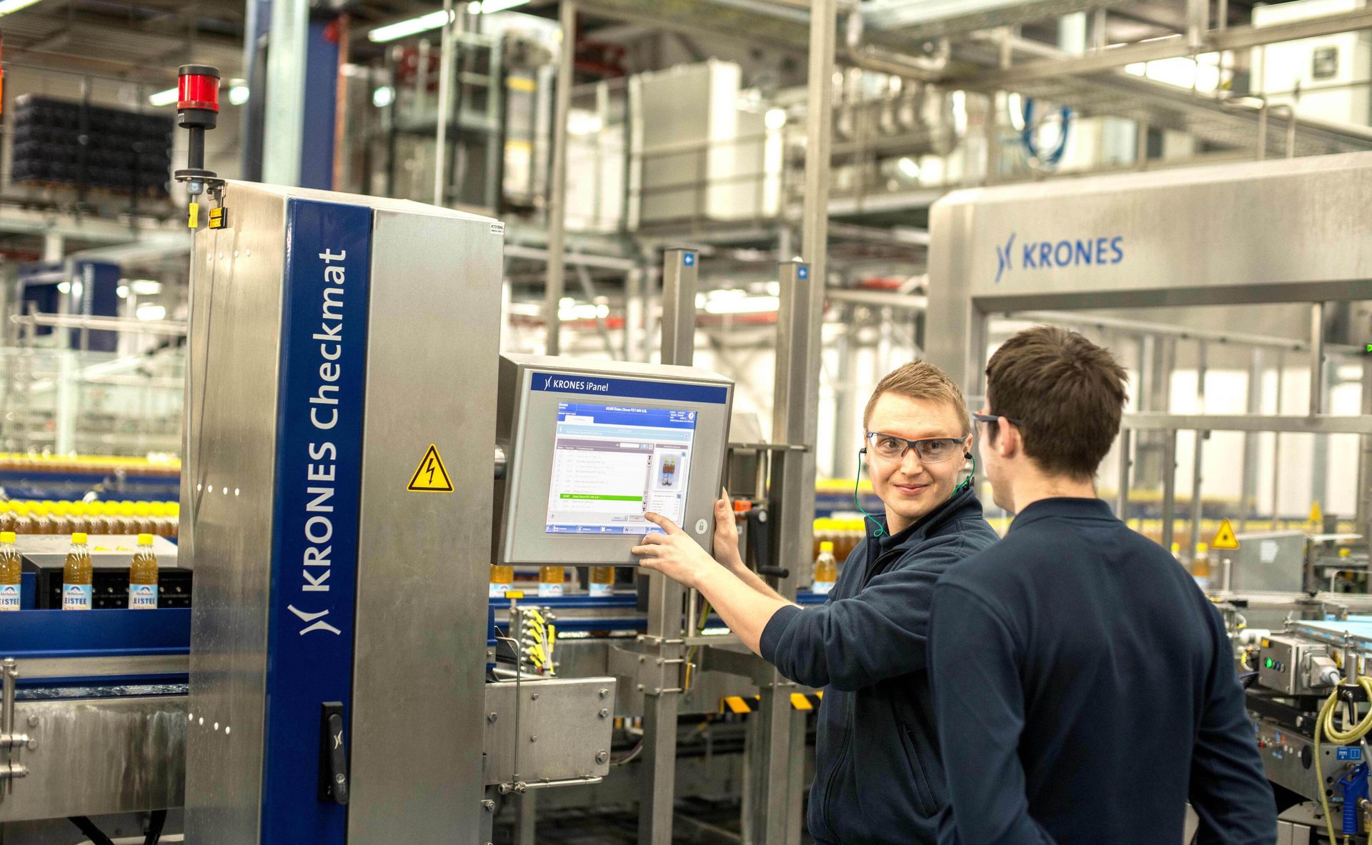Two men operate KRONES Checkmat bottling machinery in a factory.