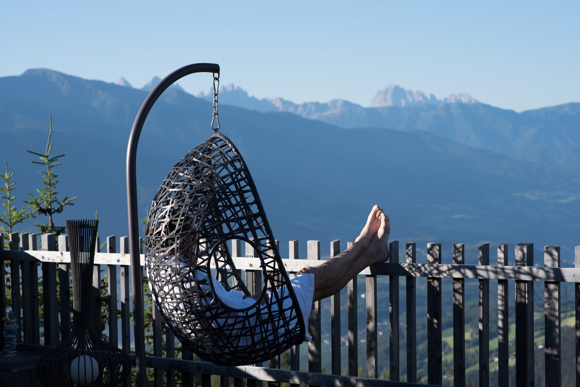 Person relaxing barefoot in a hanging chair on a balcony overlooking a mountain range.
