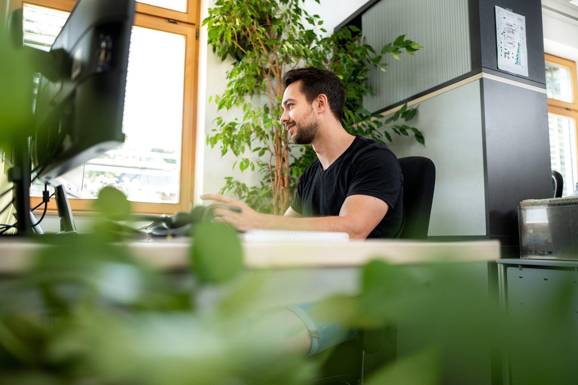 Smiling man working at a computer in an office with plants.