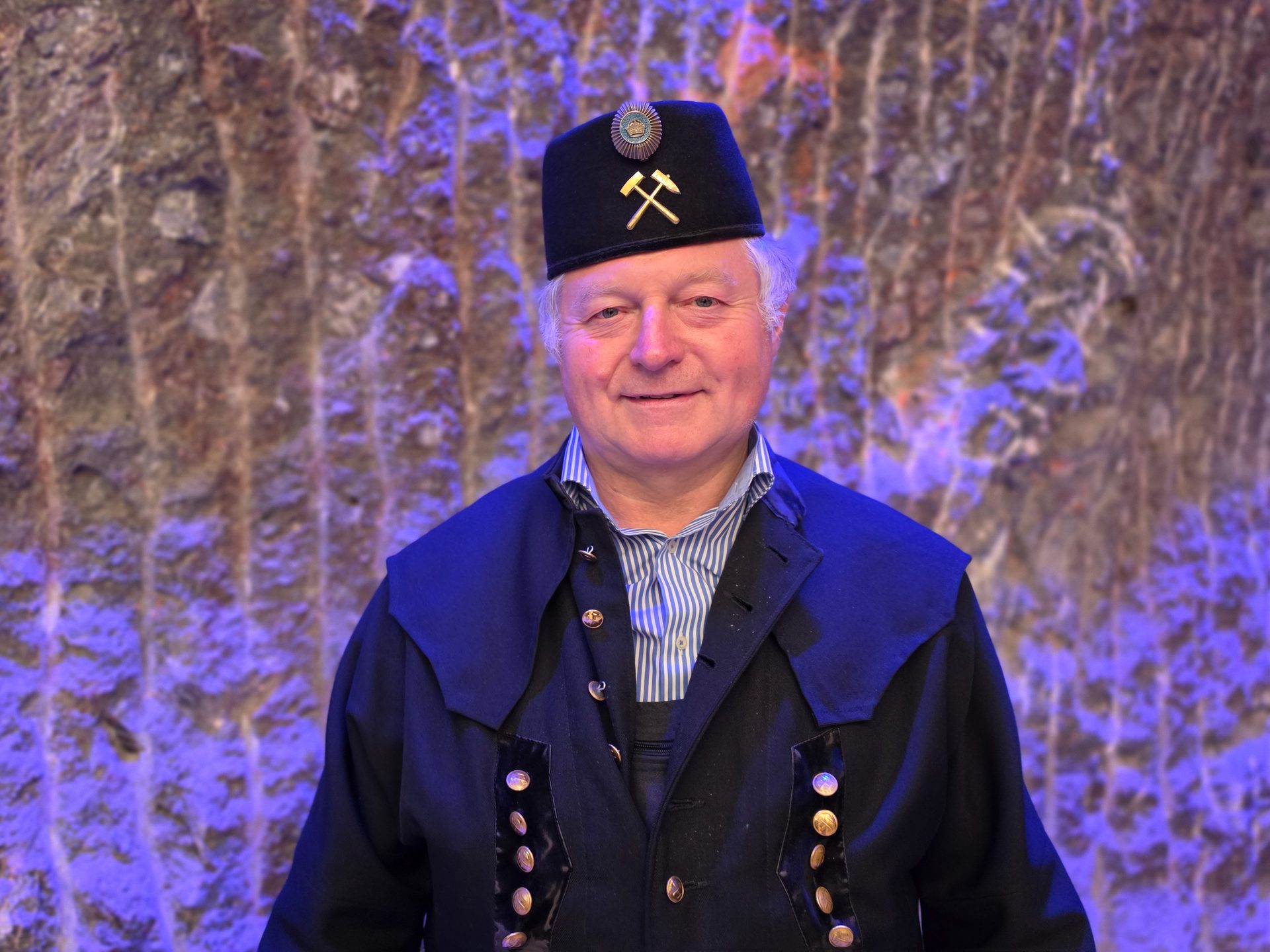 Man in traditional German mining uniform with a hat adorned with crossed hammers, smiling against a rocky background.