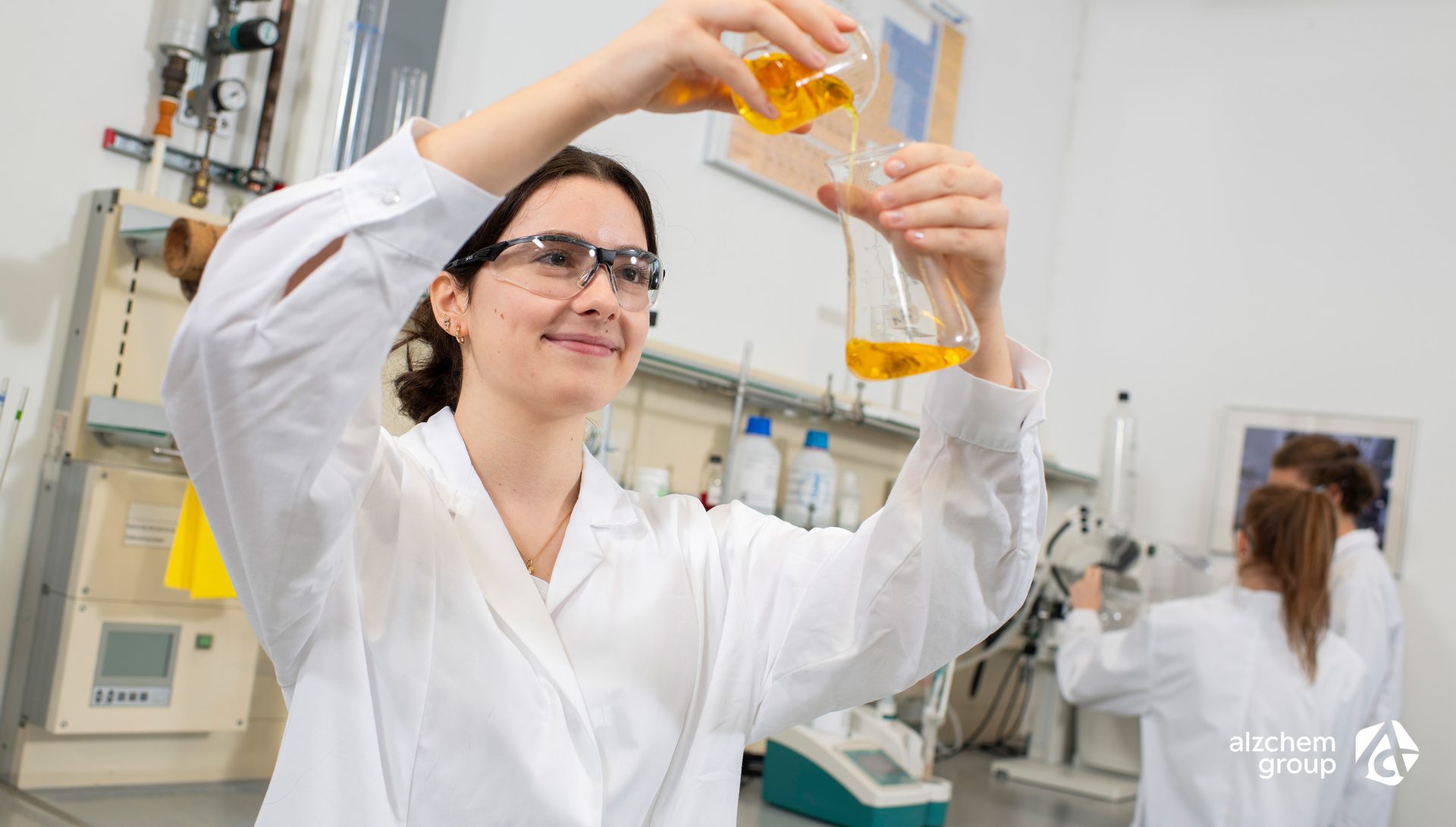 Young woman in lab coat and glasses pours yellow liquid.