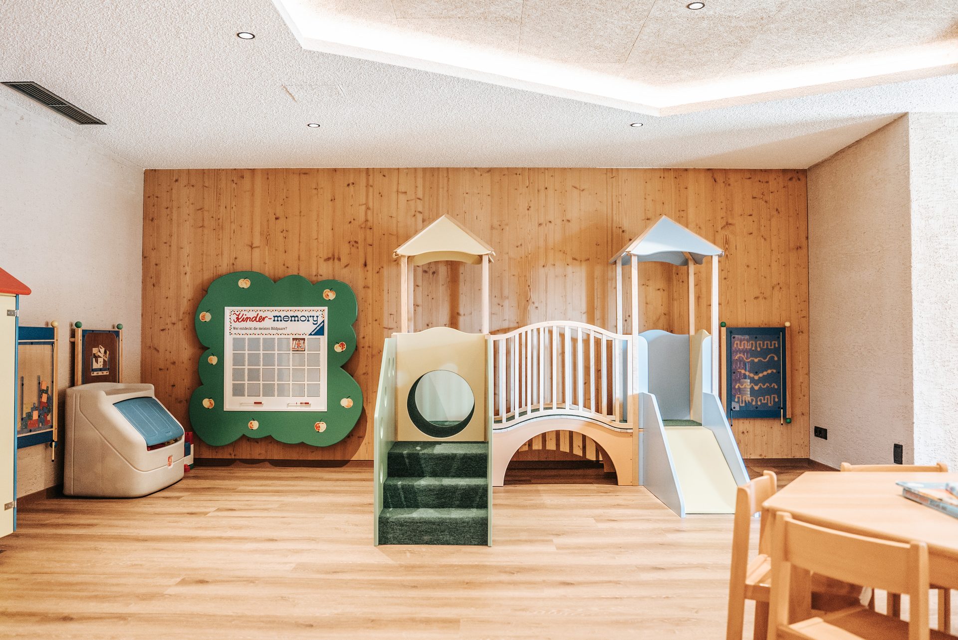 A children's playroom with a wooden climbing structure, a memory board, and a small table with chairs.