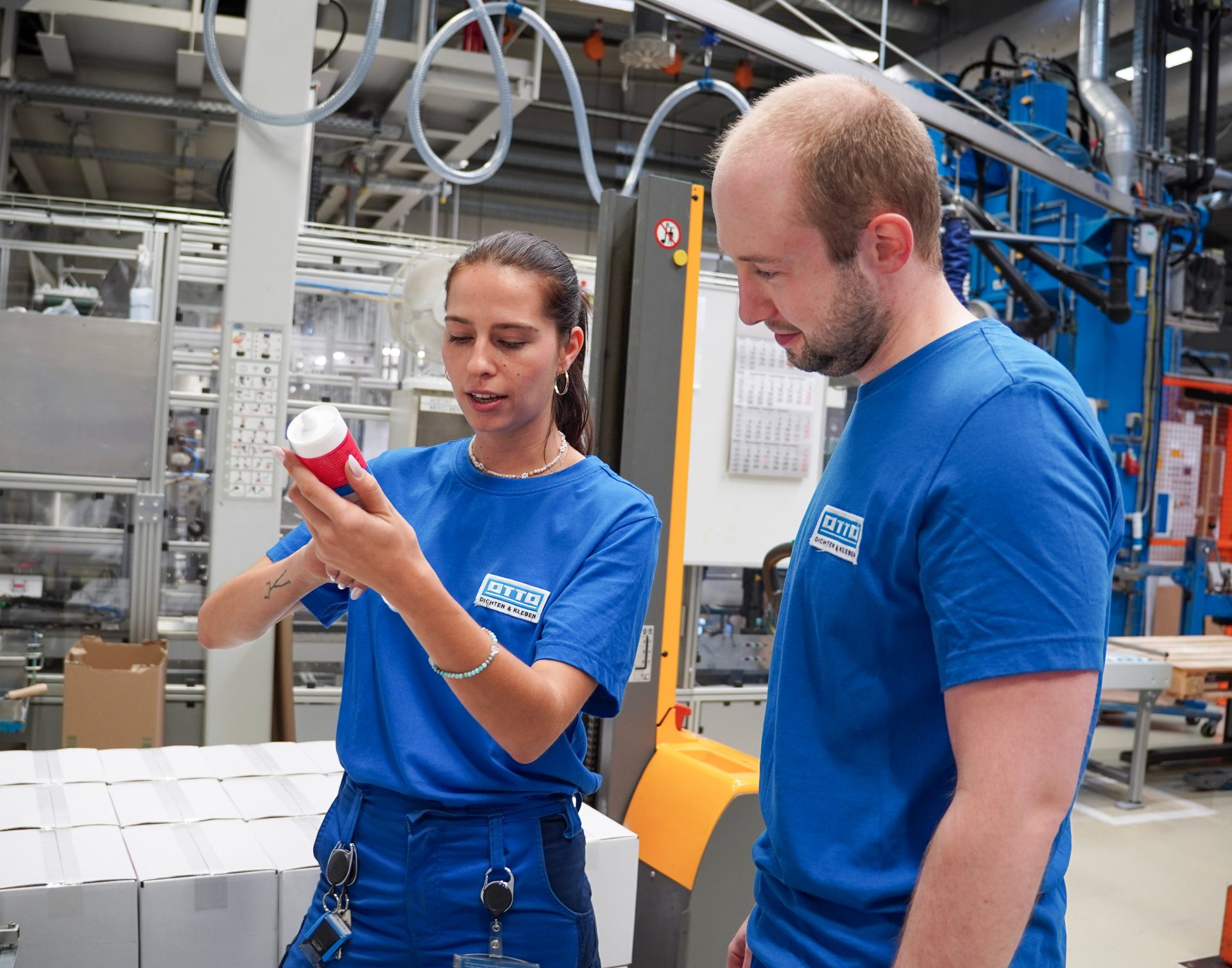 Two factory workers in blue uniforms inspecting a product tube on the manufacturing floor.